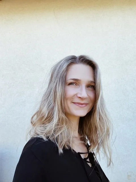 A woman with wavy blonde hair smiling in front of a plain light-colored wall.