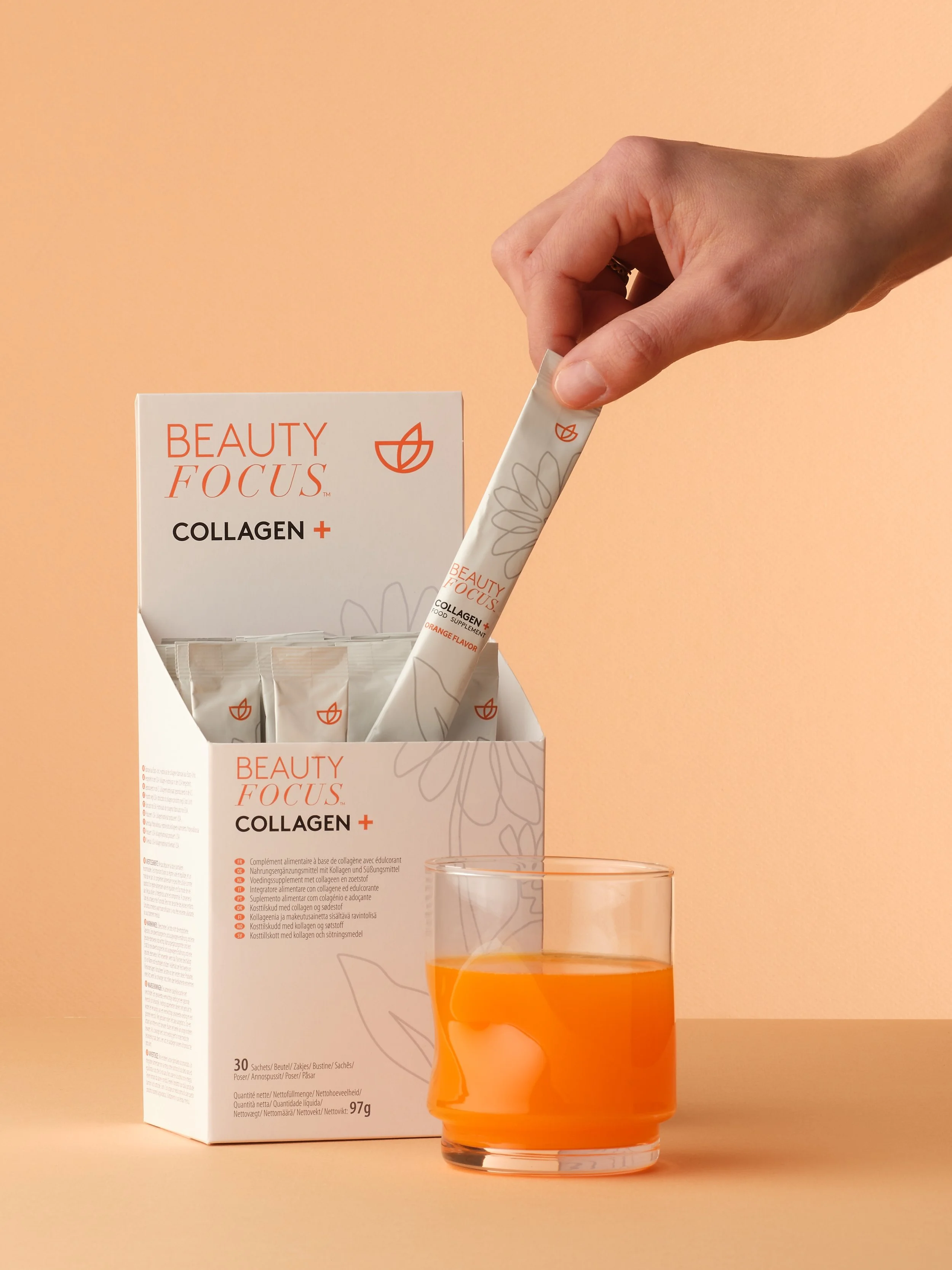 A hand holding a packaged collagen supplement stick above a glass of orange-colored liquid, with a box of collagen supplements in the background on a peach-colored surface.