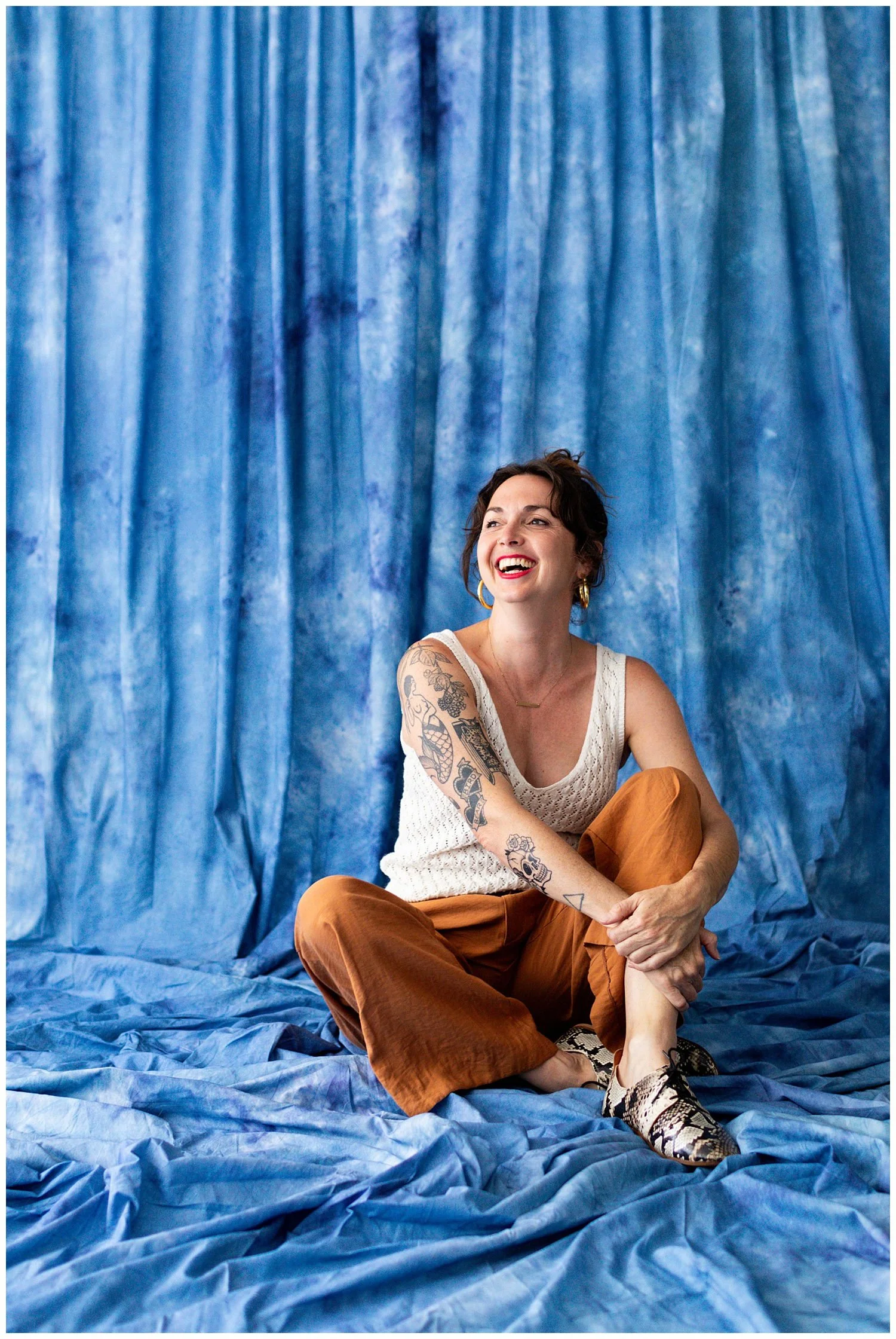 A woman with tattoos and earrings is sitting on a crumpled blue fabric backdrop, smiling and looking to her left, wearing a sleeveless white top and patterned shoes.