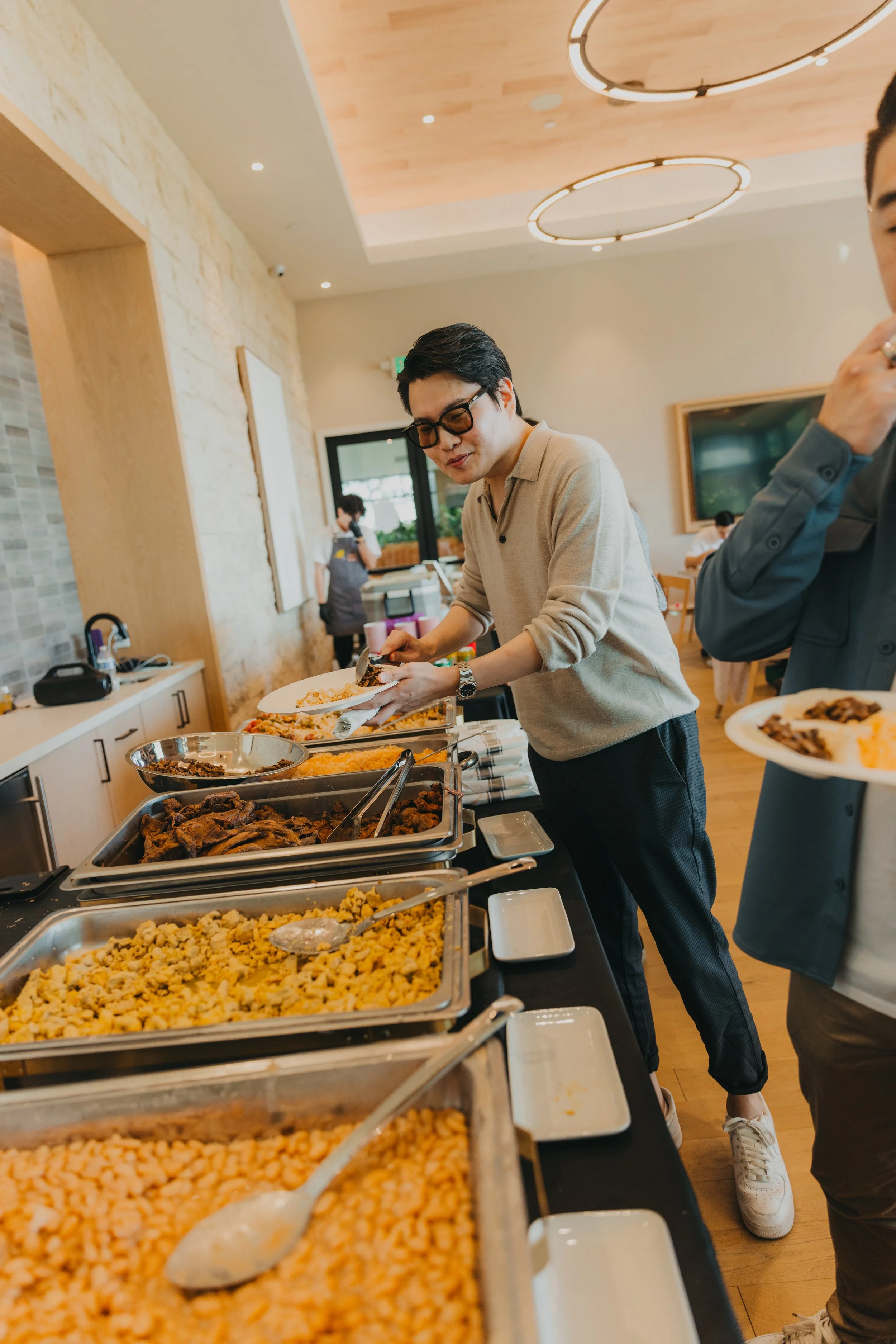 People serving themselves food at a buffet in a modern restaurant with wood-paneled ceiling and circular lighting fixtures.
