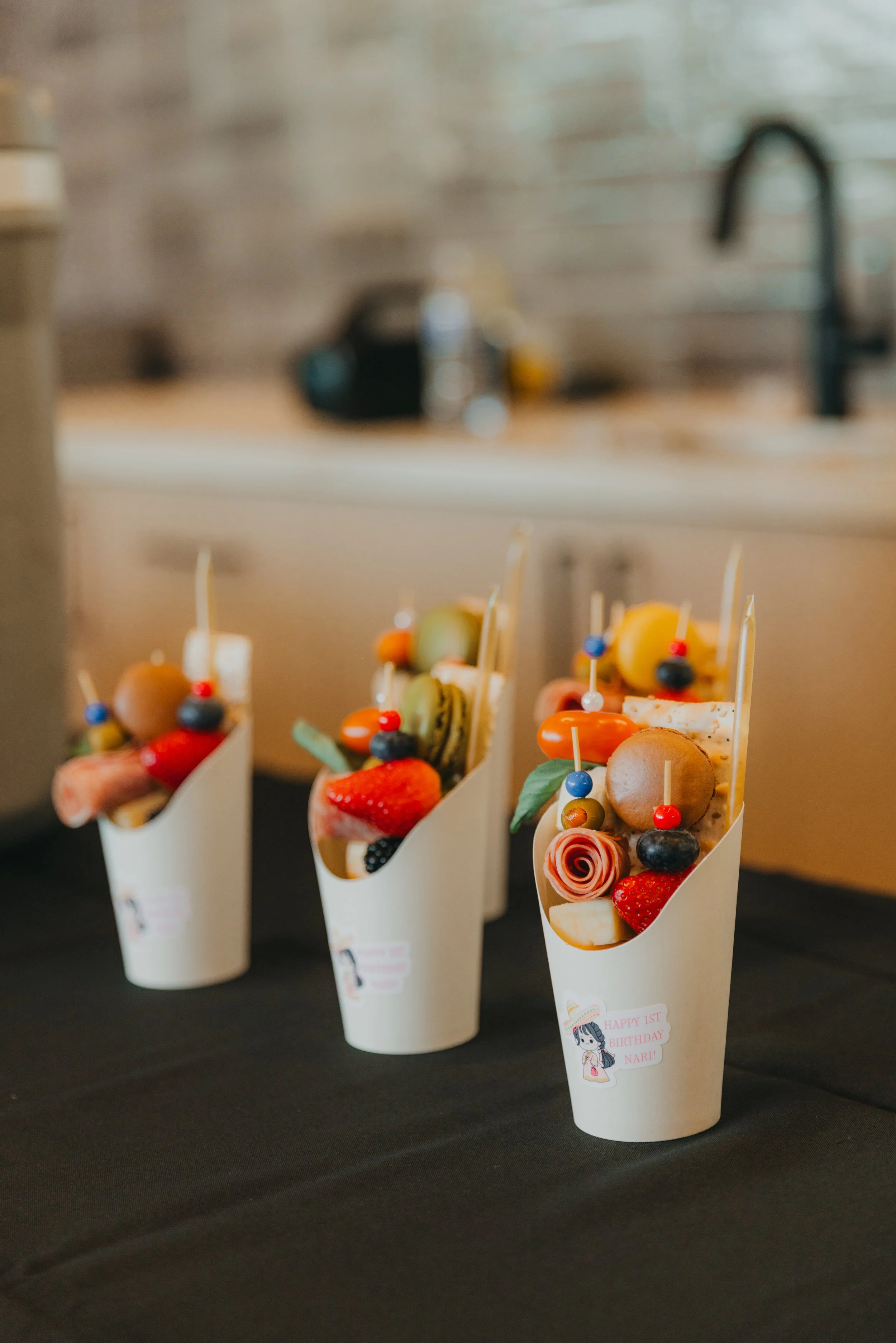 Three white cones filled with assorted candies, strawberries, macarons, and decorative elements, placed on a black surface for a celebration.