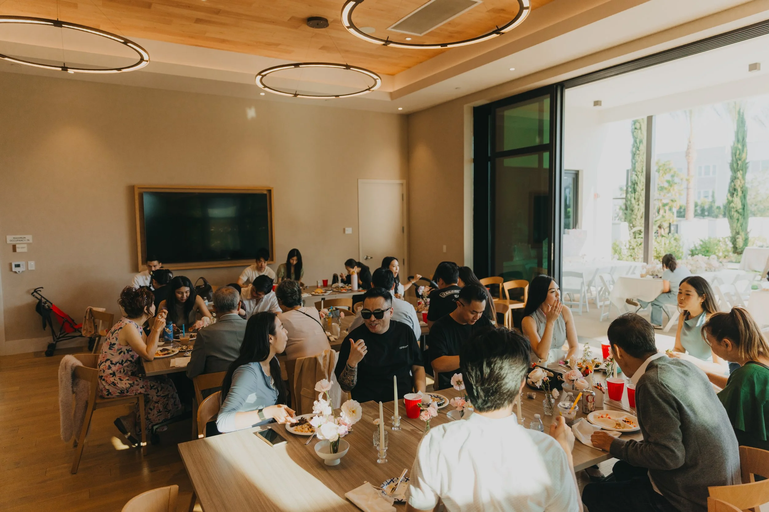 A group of people gathered around a long dining table in a bright, modern room with large windows and wooden accents, celebrating together.