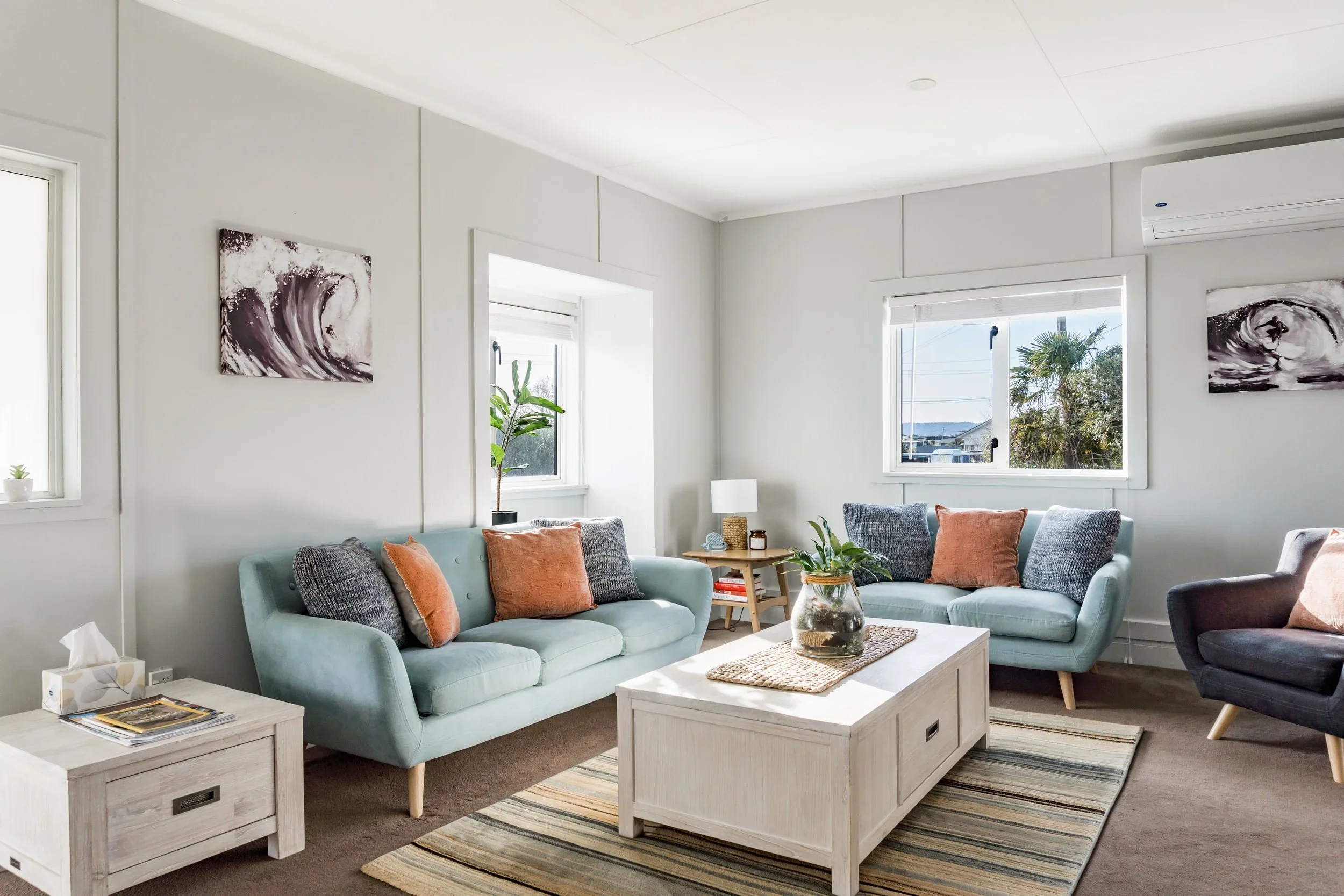 Living room with two light blue sofas, one dark gray armchair, white coffee table, side table, potted plants, abstract artwork, window with outdoor view, and neutral decor.