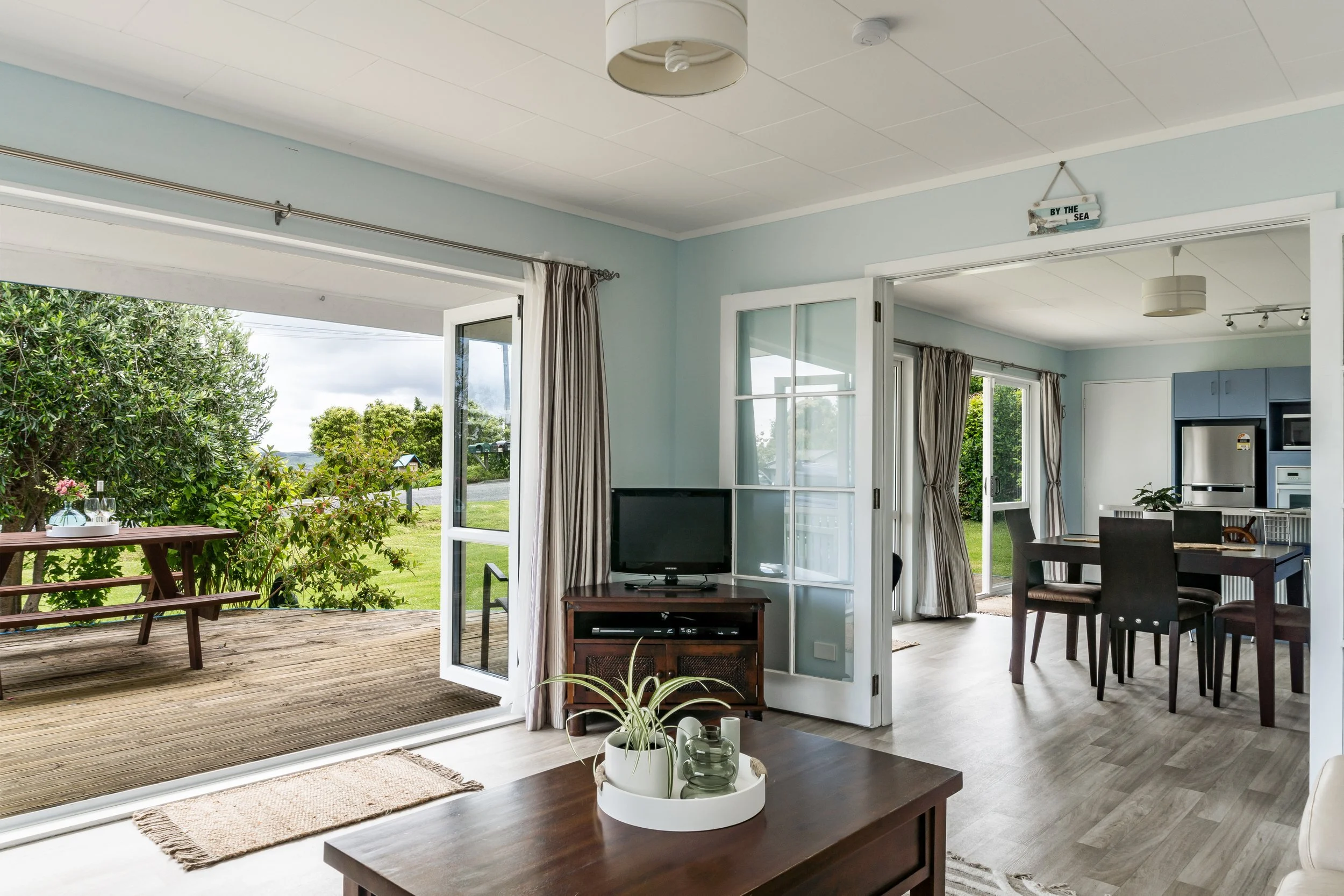 Living room with open patio door viewing green outdoor scenery, dining area with dark wood table and chairs, and kitchen in background, decorated with light blue walls and beige curtains.