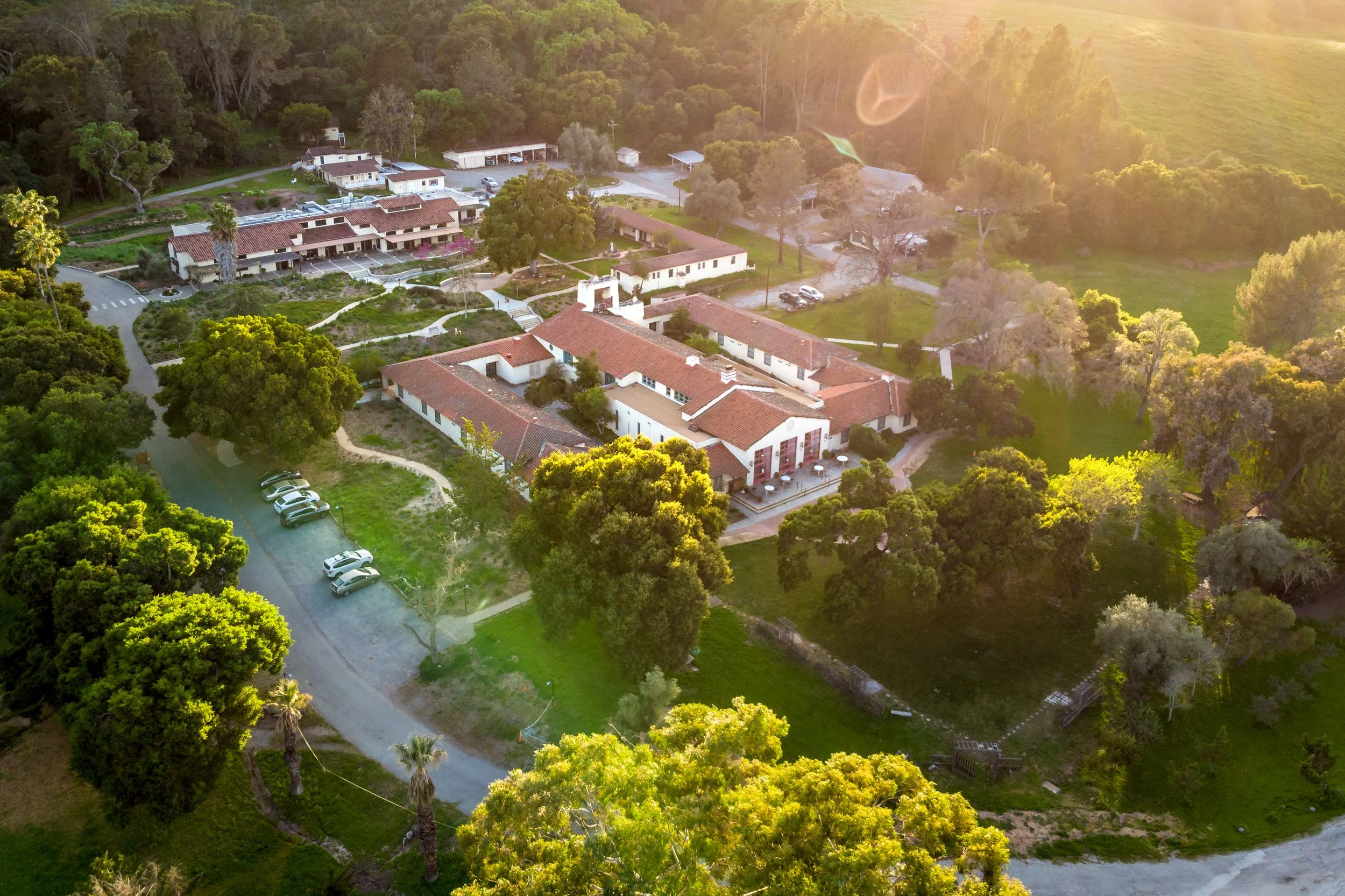 Aerial view of a small retreat center in California with red-tiled roofs surrounded by greenery and trees, with parking lots and a winding road, bathed in warm sunlight.