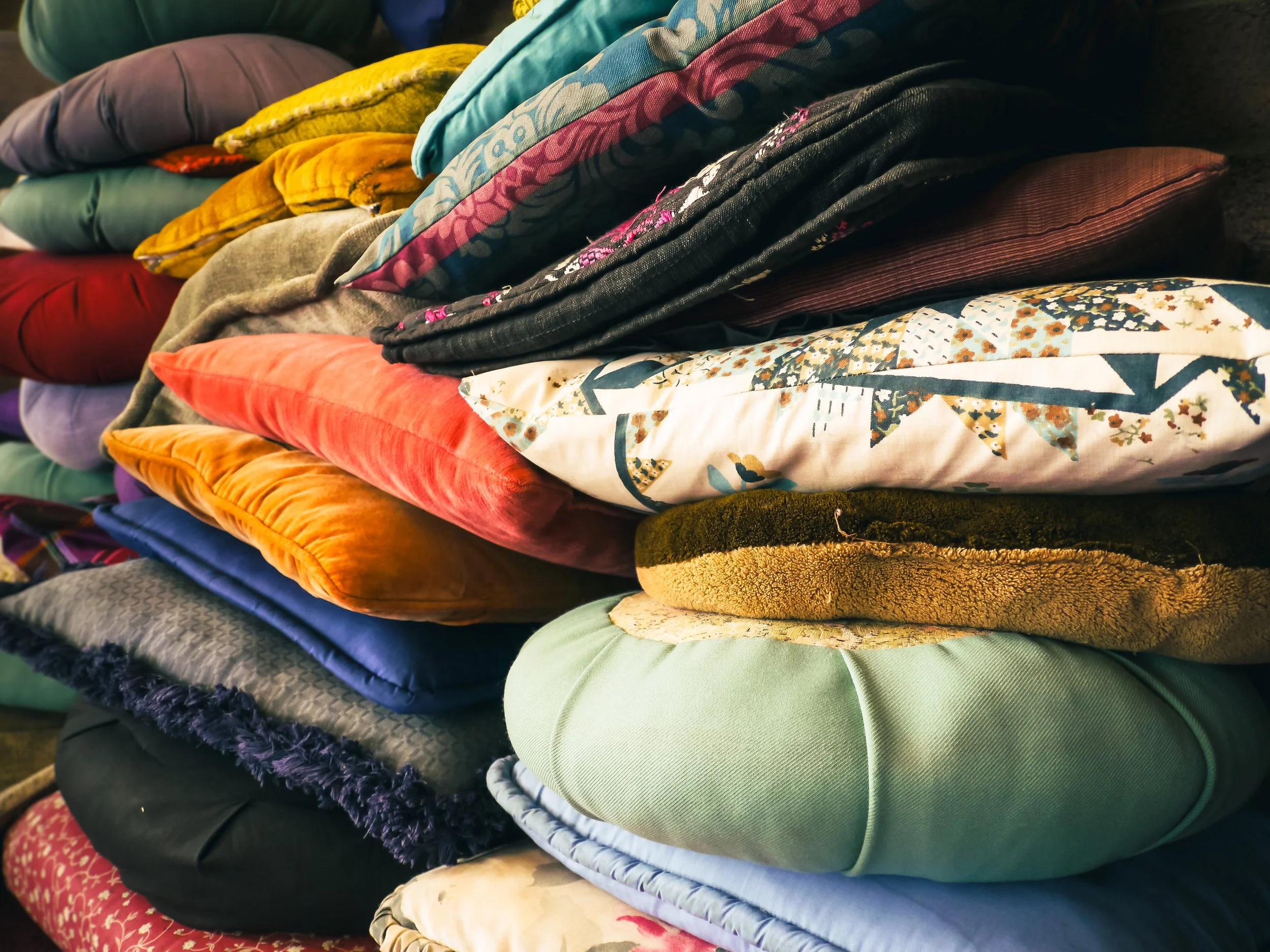 Stacked colorful cushions and pillows with various patterns, textures, and colors including green, orange, yellow, purple, black, and floral designs.