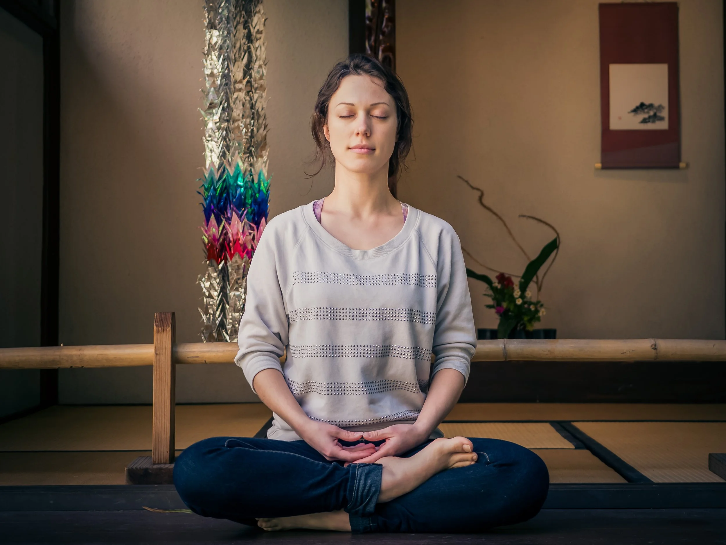 A woman practicing meditation indoors, seated cross-legged with her eyes closed and hands resting on her lap, in front of a wooden railing and a decorative Japanese scroll on the wall behind her.