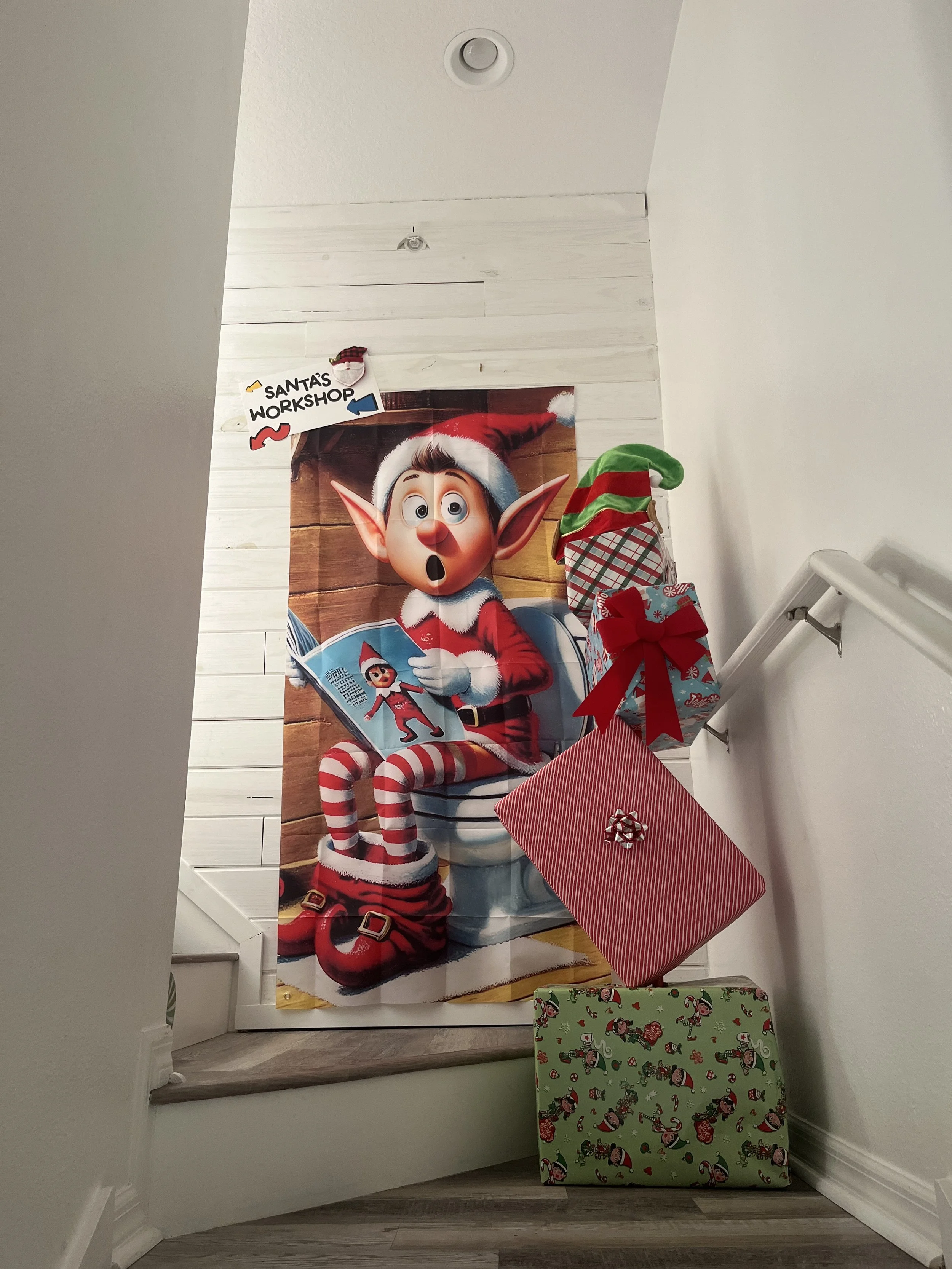 Holiday decorations including a large poster of an elf reading a book, greeting cards, and wrapped presents, on a staircase landing with white walls.
