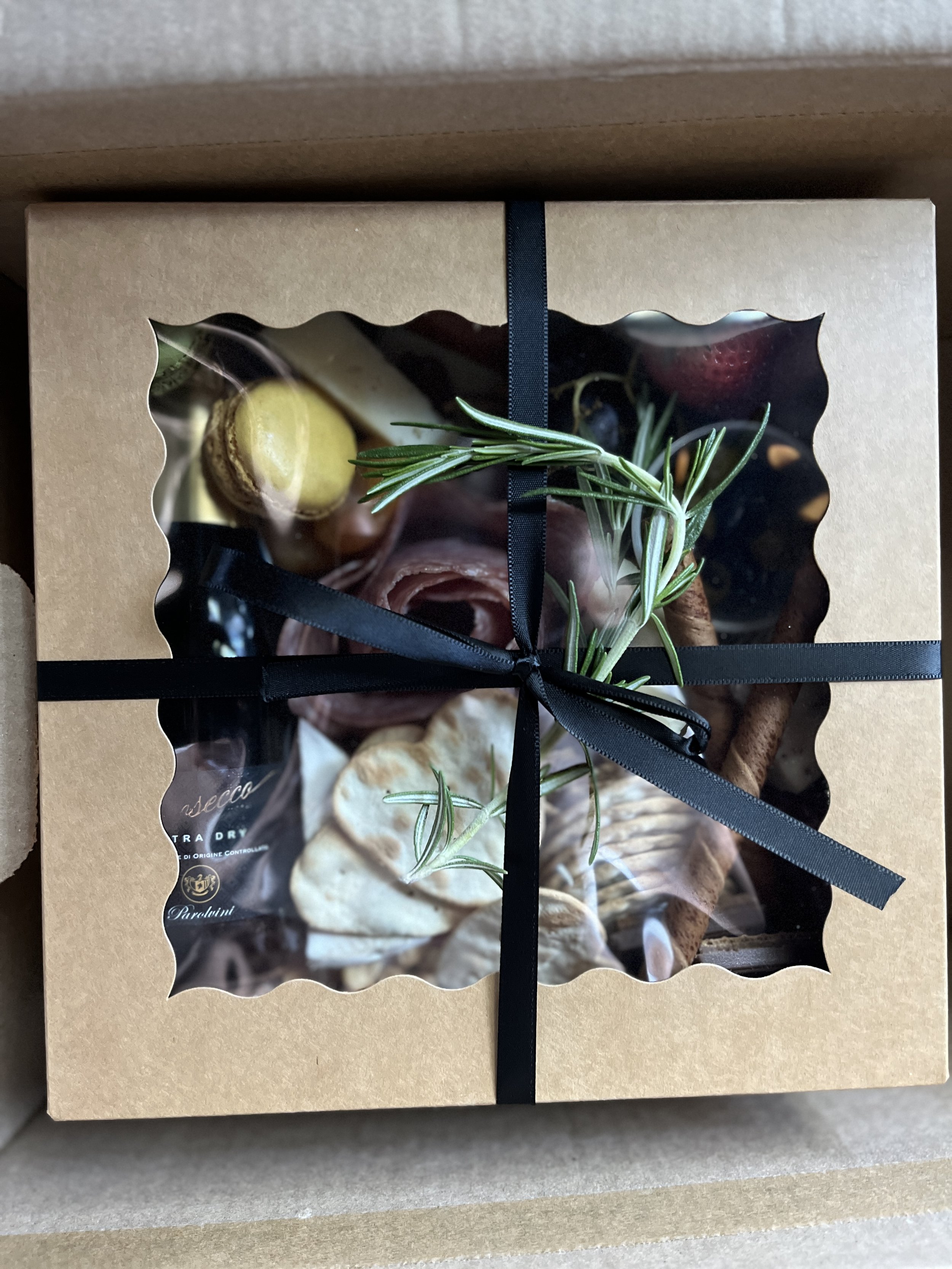 A gift box with a clear window showing assorted food items, including slices of meat, bread, olives, and garnished with rosemary, wrapped with a black ribbon.