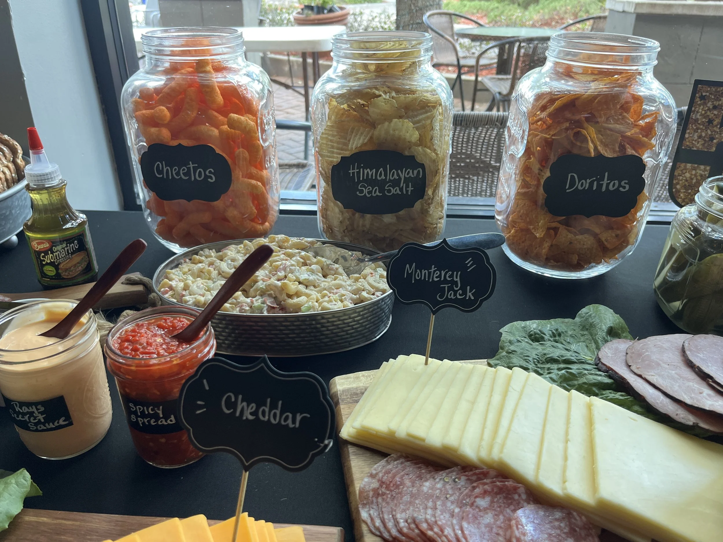 A spread of snacks and condiments on a black table, including jars of Cheetos, Himalayan Sea Salt, Doritos, a bowl of macaroni salad, slices of cheddar cheese, deli meats, and sauces, with a window and patio furniture in the background.