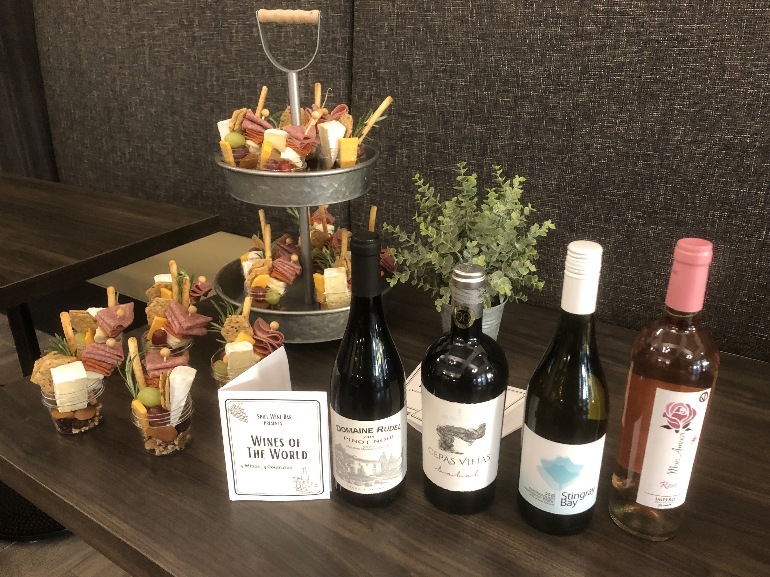 A display of four bottles of wine and several small appetizers on a dark wooden table, with a small potted plant and a two-tiered tray of assorted finger foods in the background.