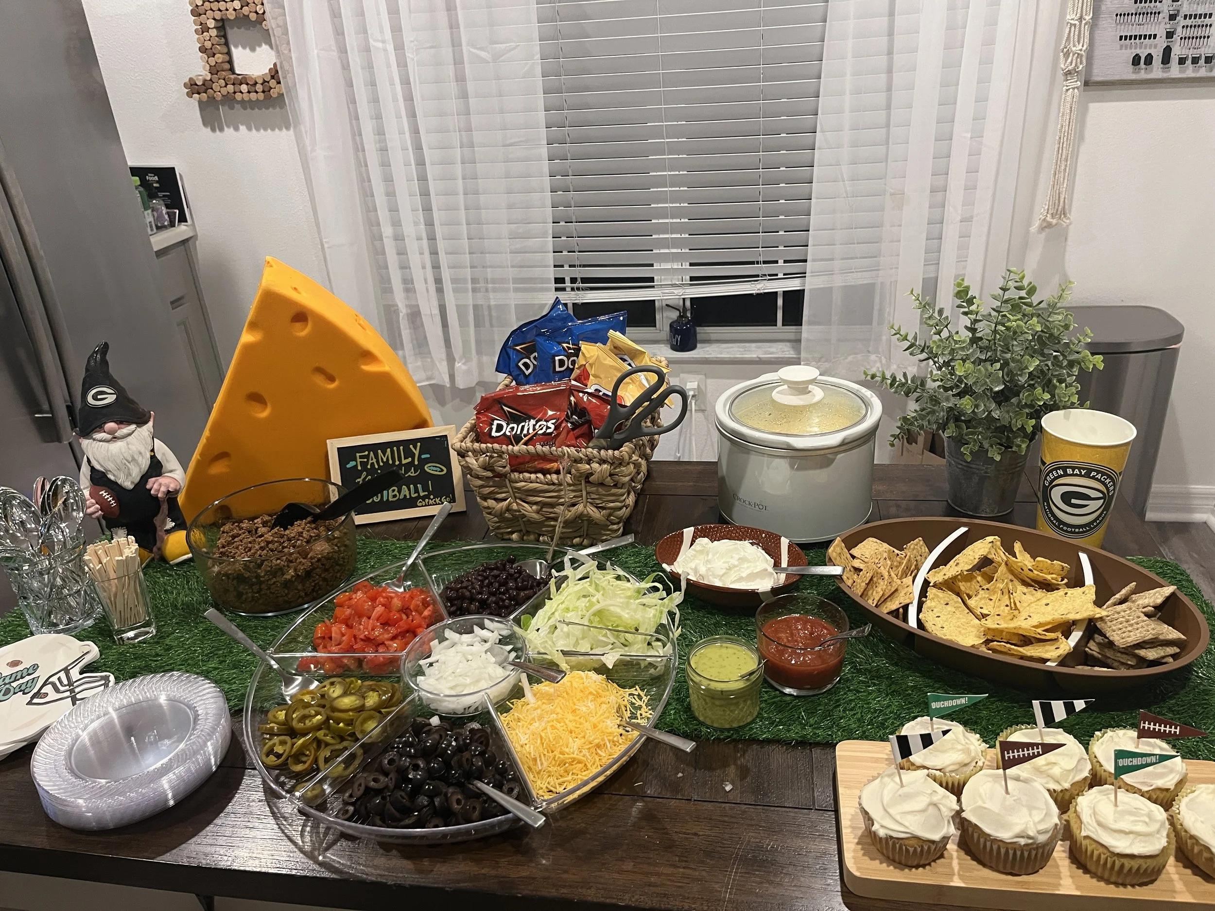 A table set for a family football gathering with chips, salsa, guacamole, black olives, shredded cheese, chopped tomatoes, chopped onions, jalapenos, sour cream, and cupcakes with sports-themed flags. There are snack bags, a slow cooker, a plant, and
