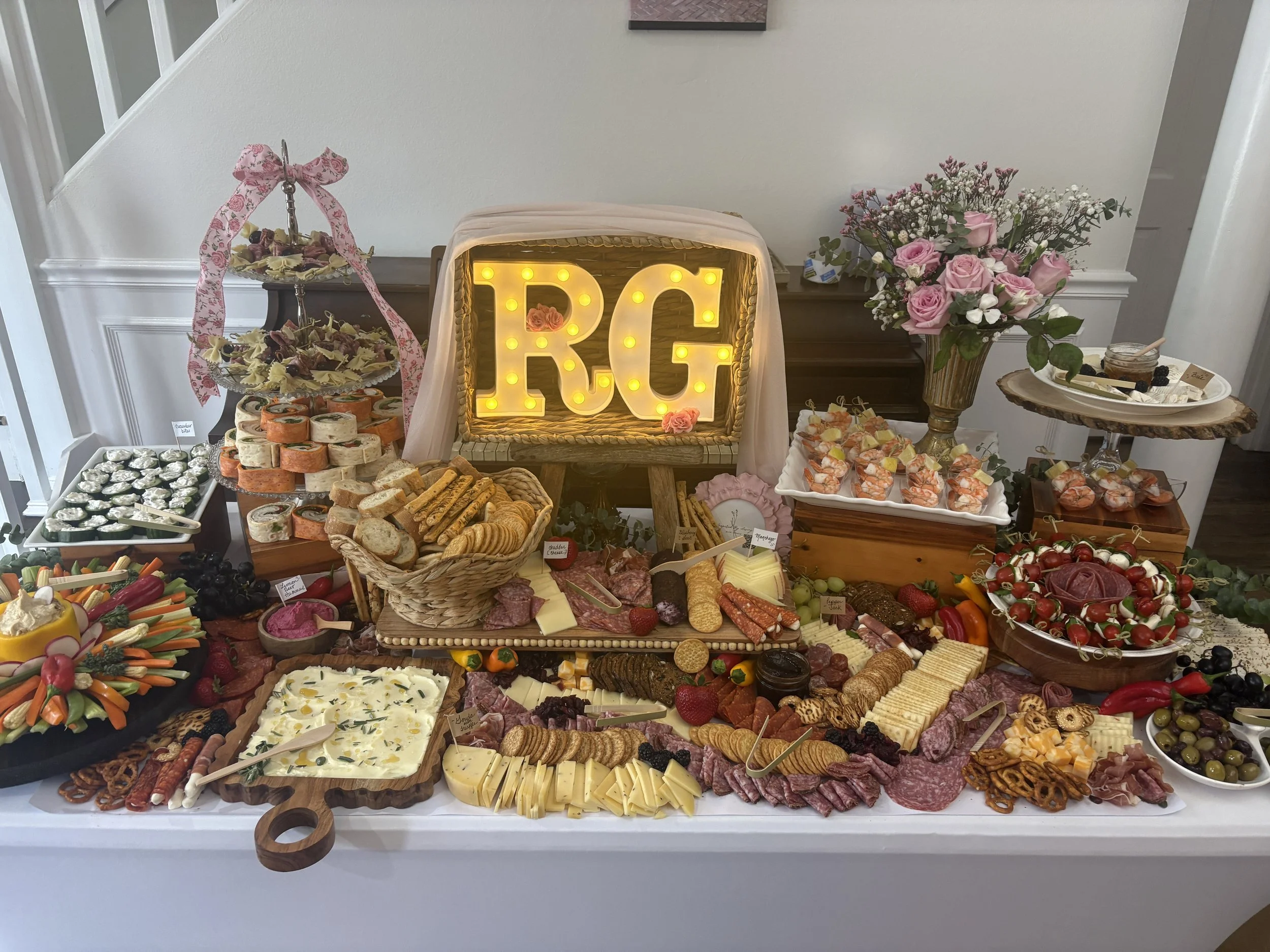 A large table with a variety of cheeses, meats, vegetables, fruits, and crackers, arranged as a charcuterie board for a celebration. In the background, there is a decorative sign with the word "LOVE" in illuminated letter lights, alongside pink roses