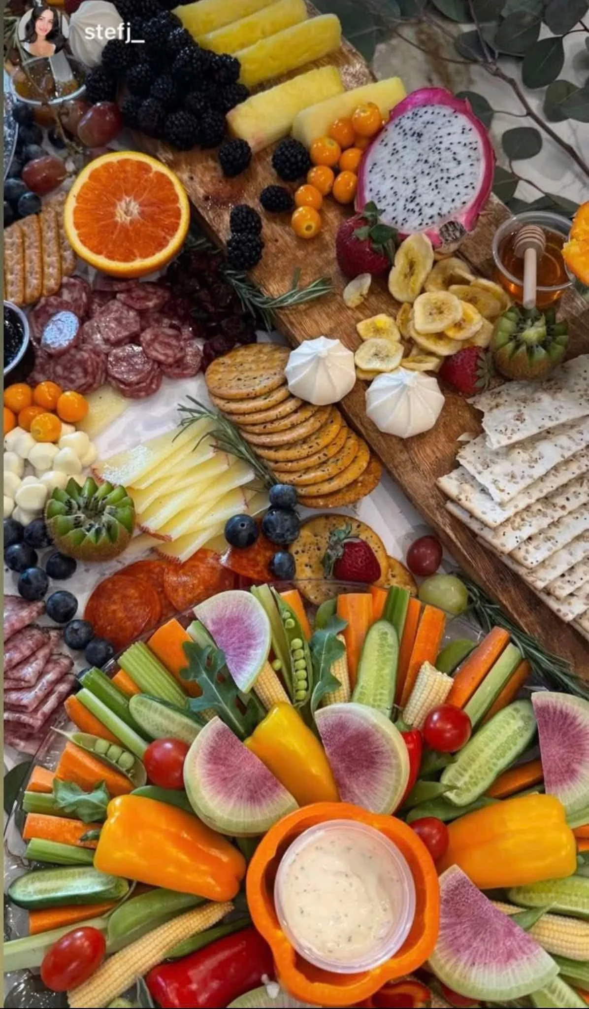 A colorful assortment of fresh fruits, vegetables, crackers, meats, cheese, and dips arranged for a buffet or appetizer platter, featuring watermelon, oranges, grapes, bananas, strawberries, blackberries, cherry tomatoes, carrots, celery, bell pepper