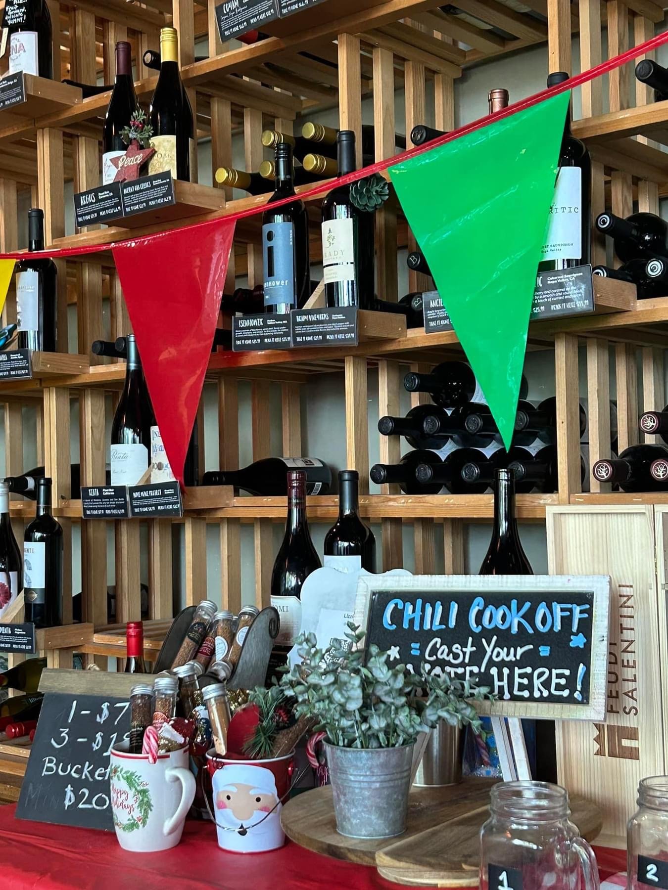 Wine bottles on wooden shelves, a sign for a chili cook-off event, and festive mugs with candy canes on a counter decorated for holiday shopping.