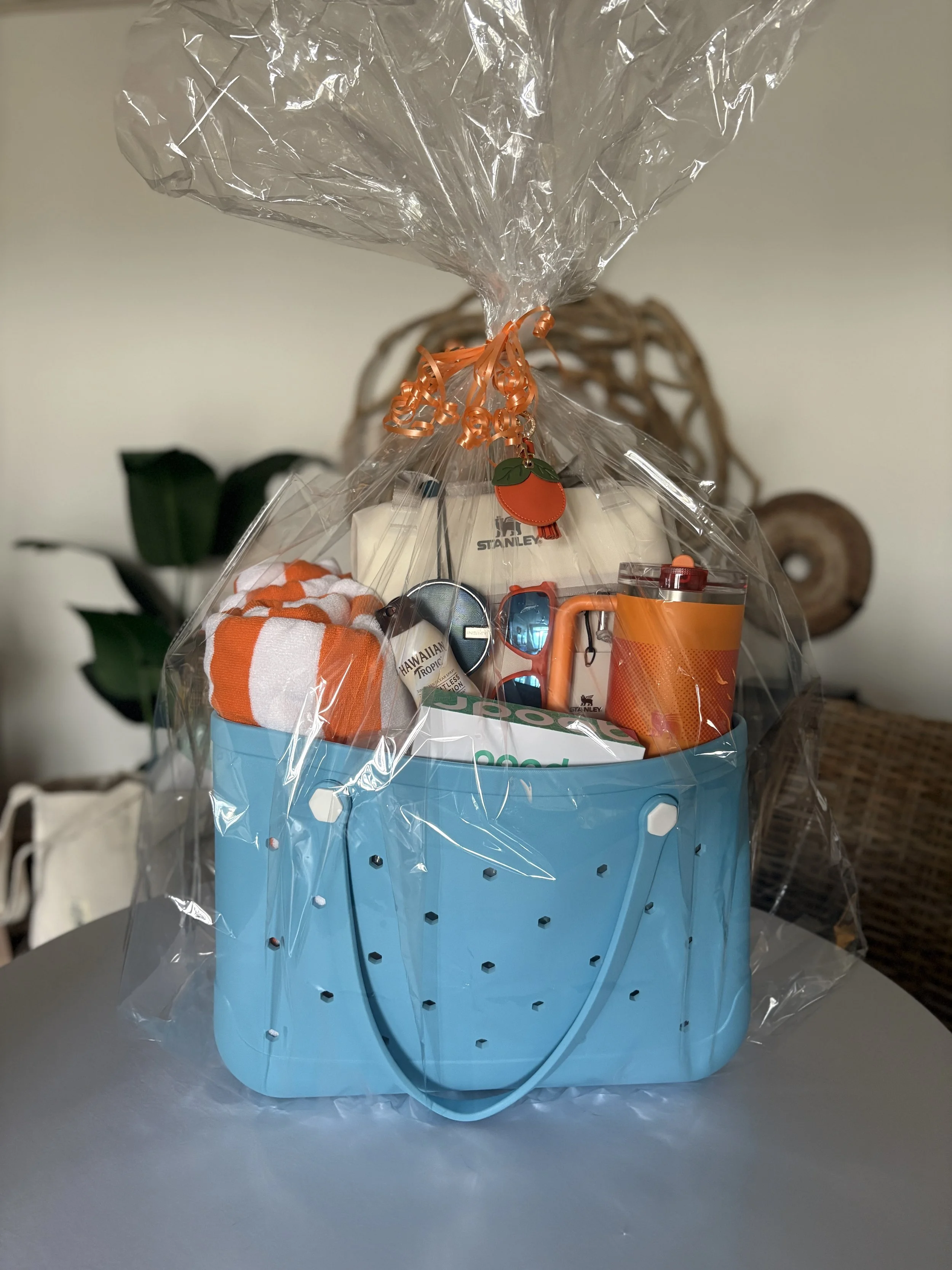 A gift basket wrapped in clear plastic with orange curly ribbon, containing a white Stanley brand object, orange and white striped towel, sunglasses, and an orange water bottle, placed on a gray surface with wicker and plant in the background.