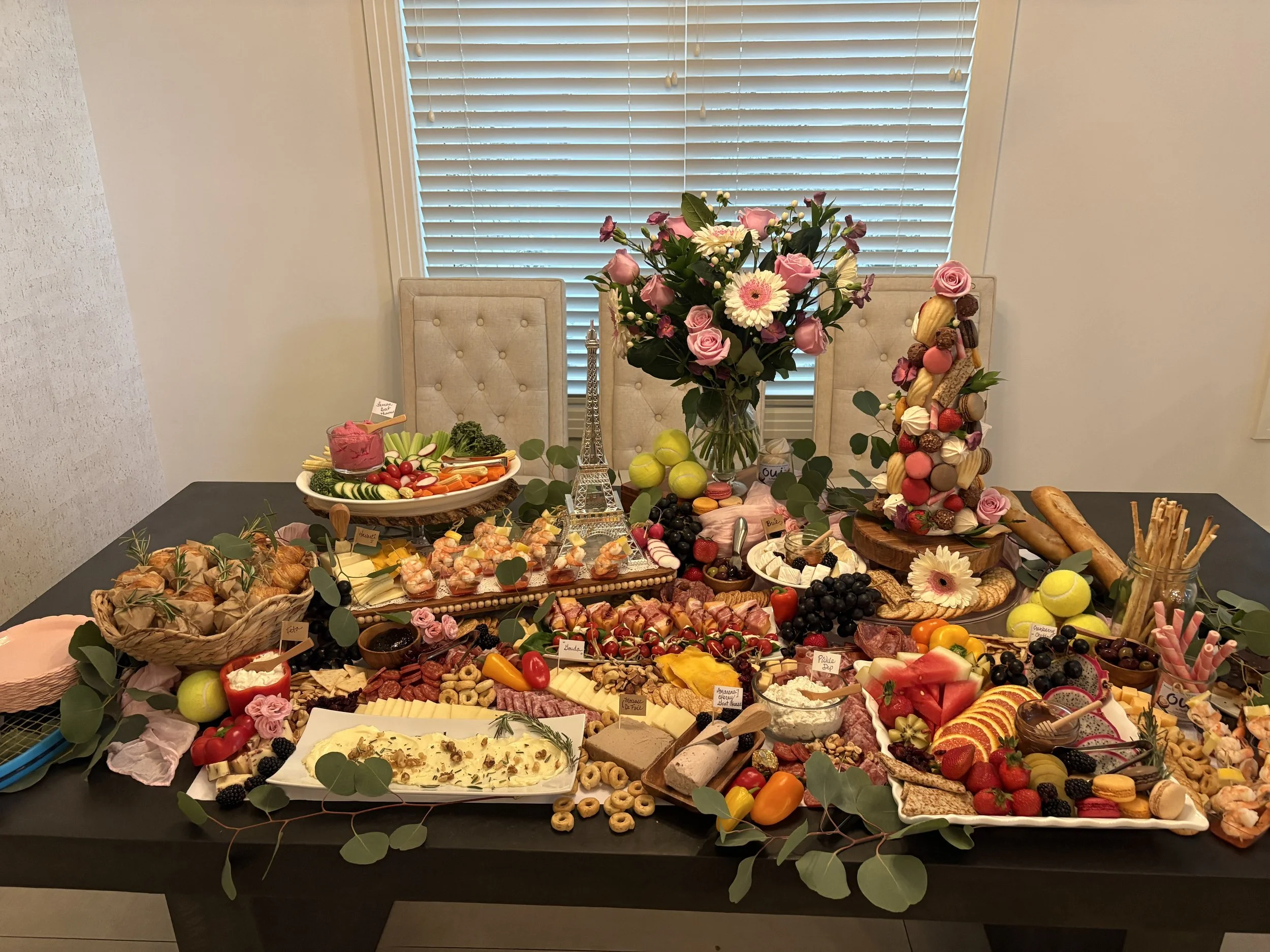 A table filled with various desserts and snacks, decorated with flowers and greenery. There is a large bouquet of pink roses and flowers in the center, with cheese, fruits, and sweets surrounding it. A small Eiffel Tower model is also on the table.