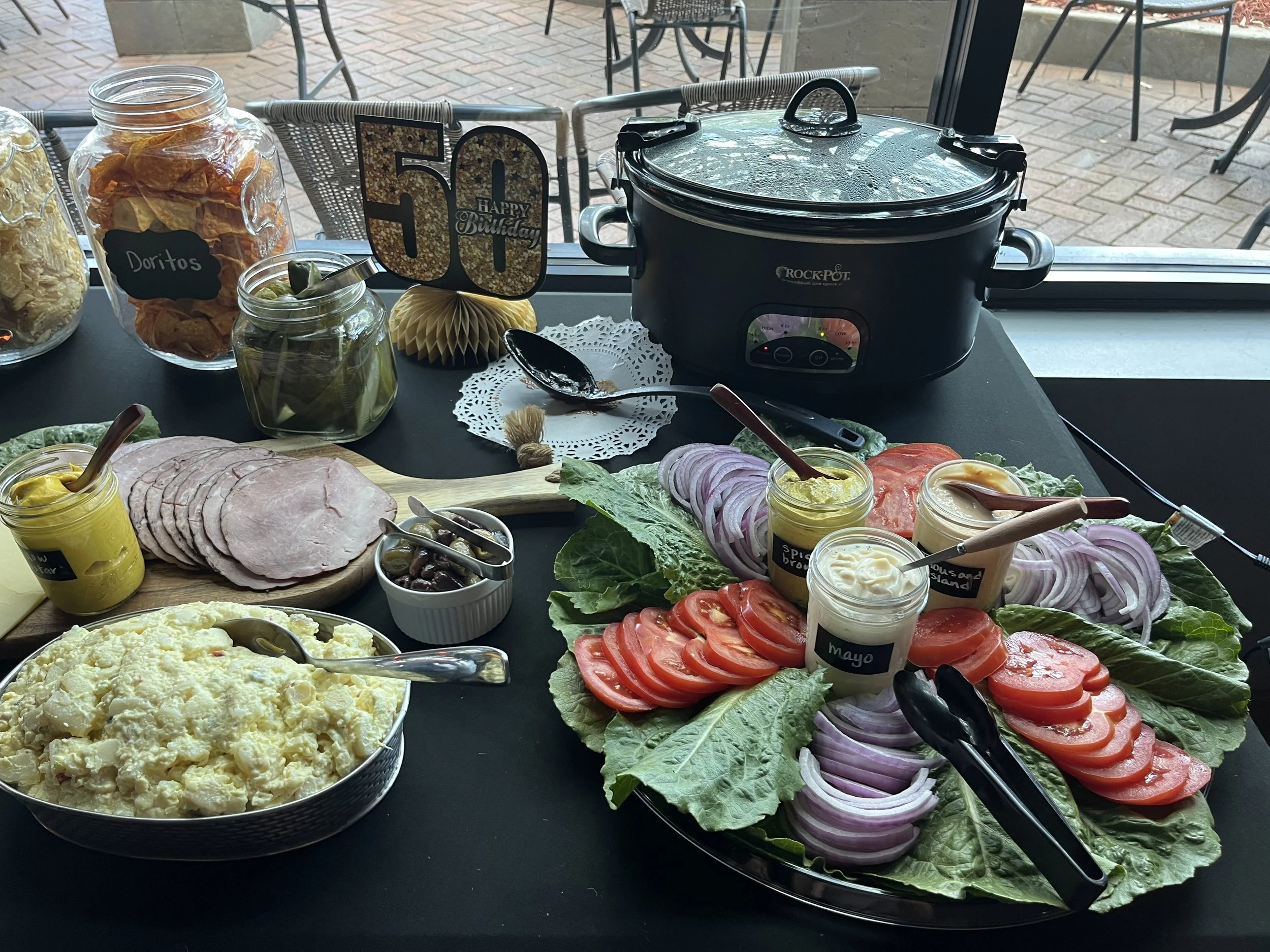 A table set with various food items including a platter of sliced tomatoes and onions, bowls of mayonnaise, potato salad, and mustard, a plate with slices of ham, condiments, a jar of pickles, chips, and a birthday sign with the number 50, a slow coo