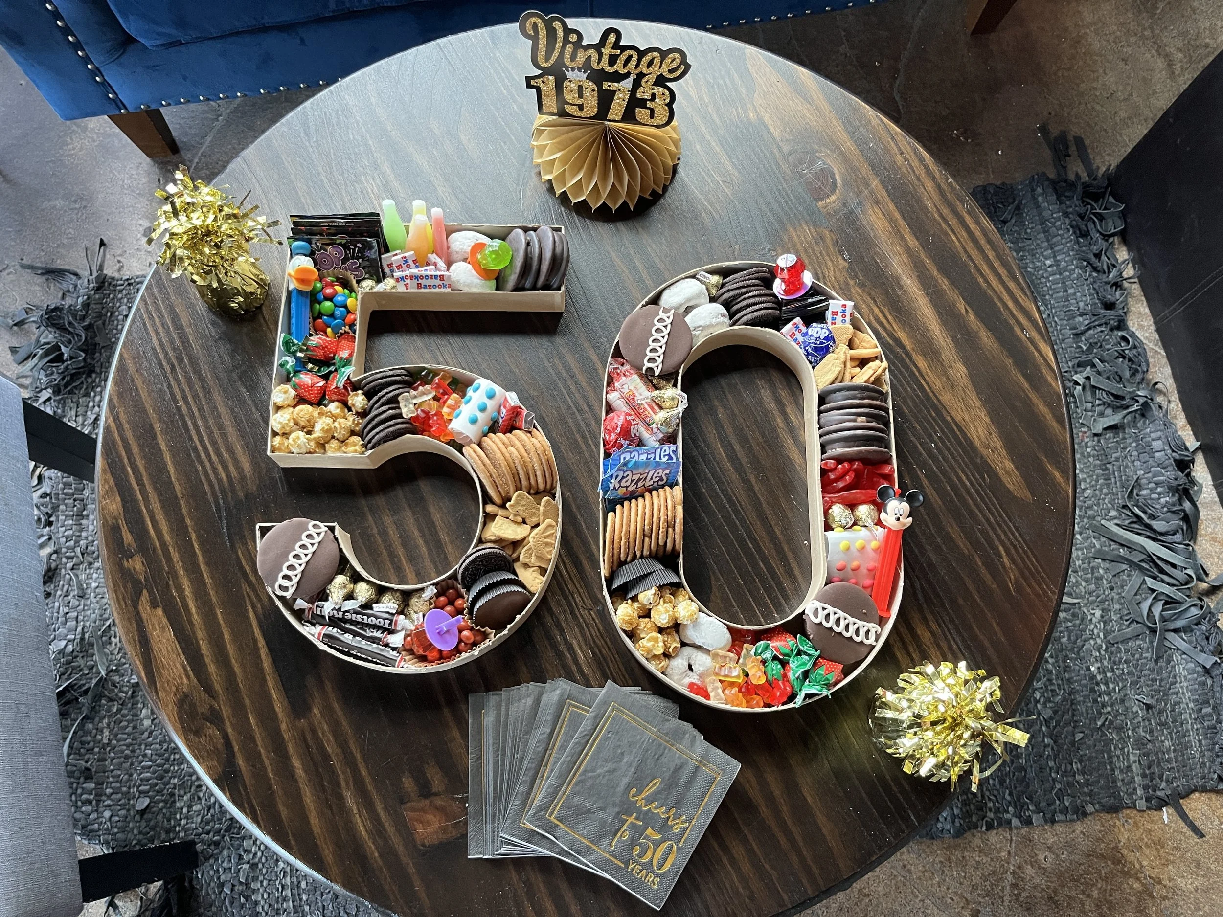 Decorative numbers '50' filled with various candies, chocolates, and snacks on a wooden table, with a sign that reads 'Vintage 1973' and a small gift bag.