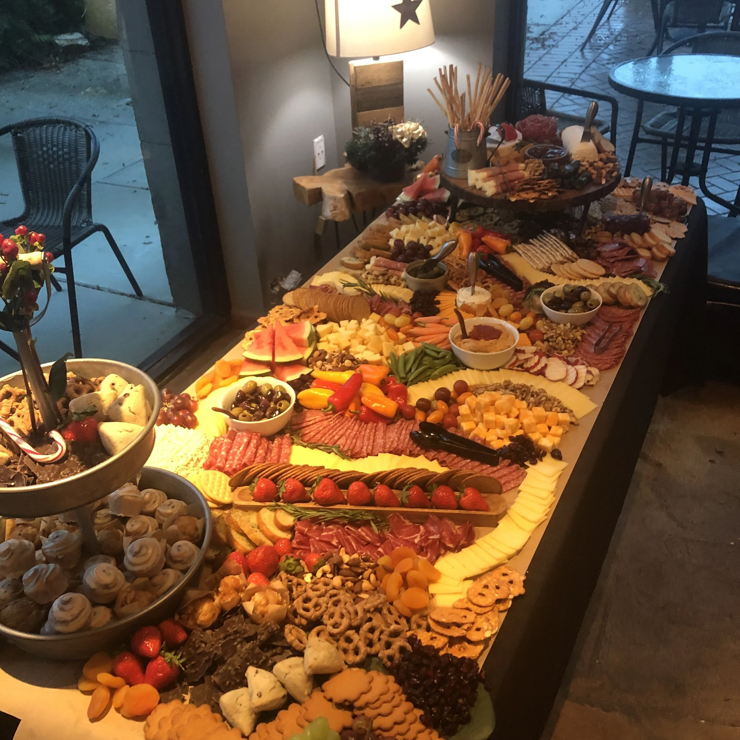 A long table set with a variety of cheeses, meats, fruits, crackers, and snacks for a buffet. Behind the table is a window showing an outdoor patio area.