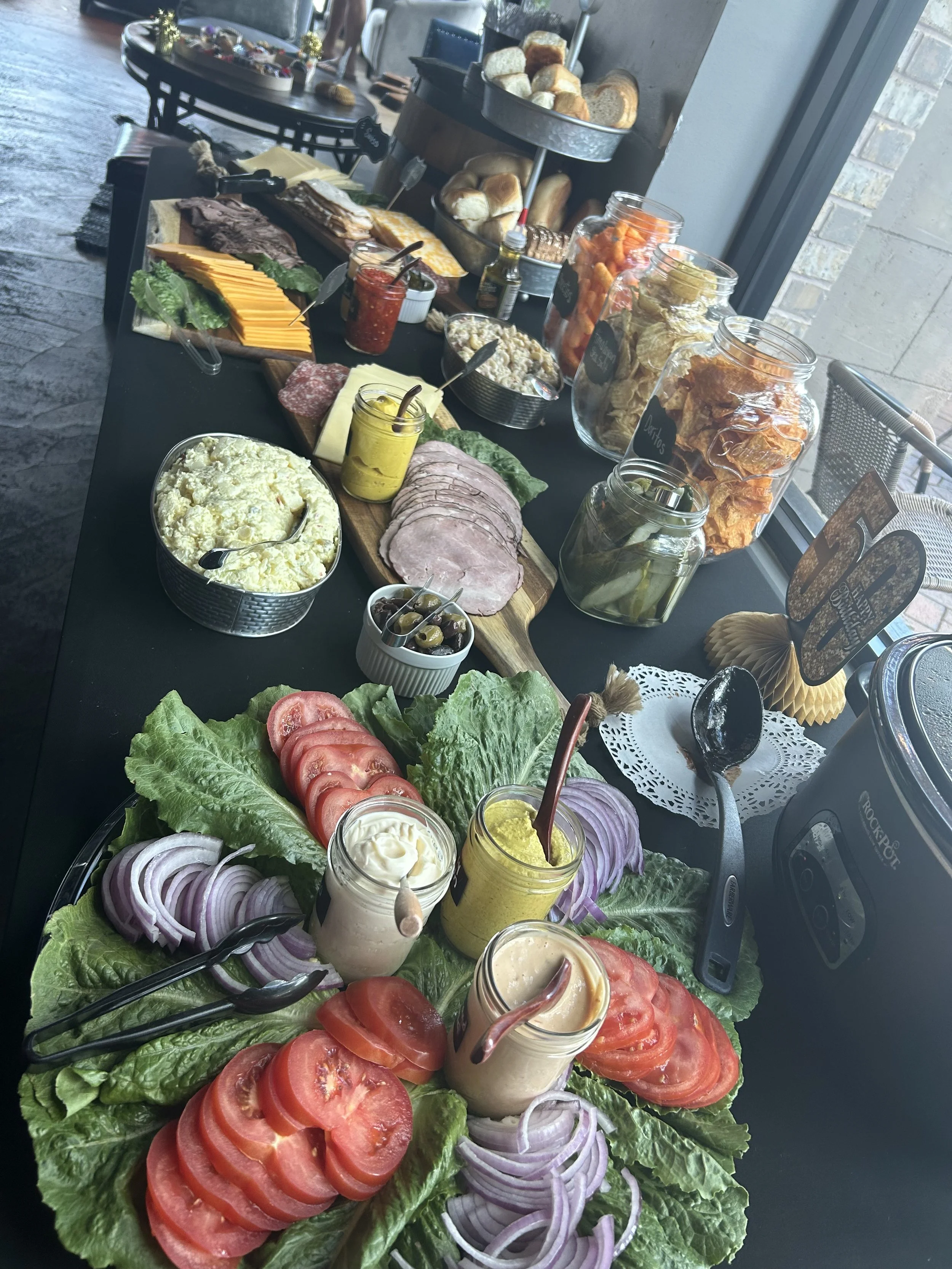 A table set with various breakfast foods, including sliced tomatoes, onions, lettuce, mayonnaise, and mustard in jars, as well as deli meats, cheeses, pickles, and an assortment of chips and bread.