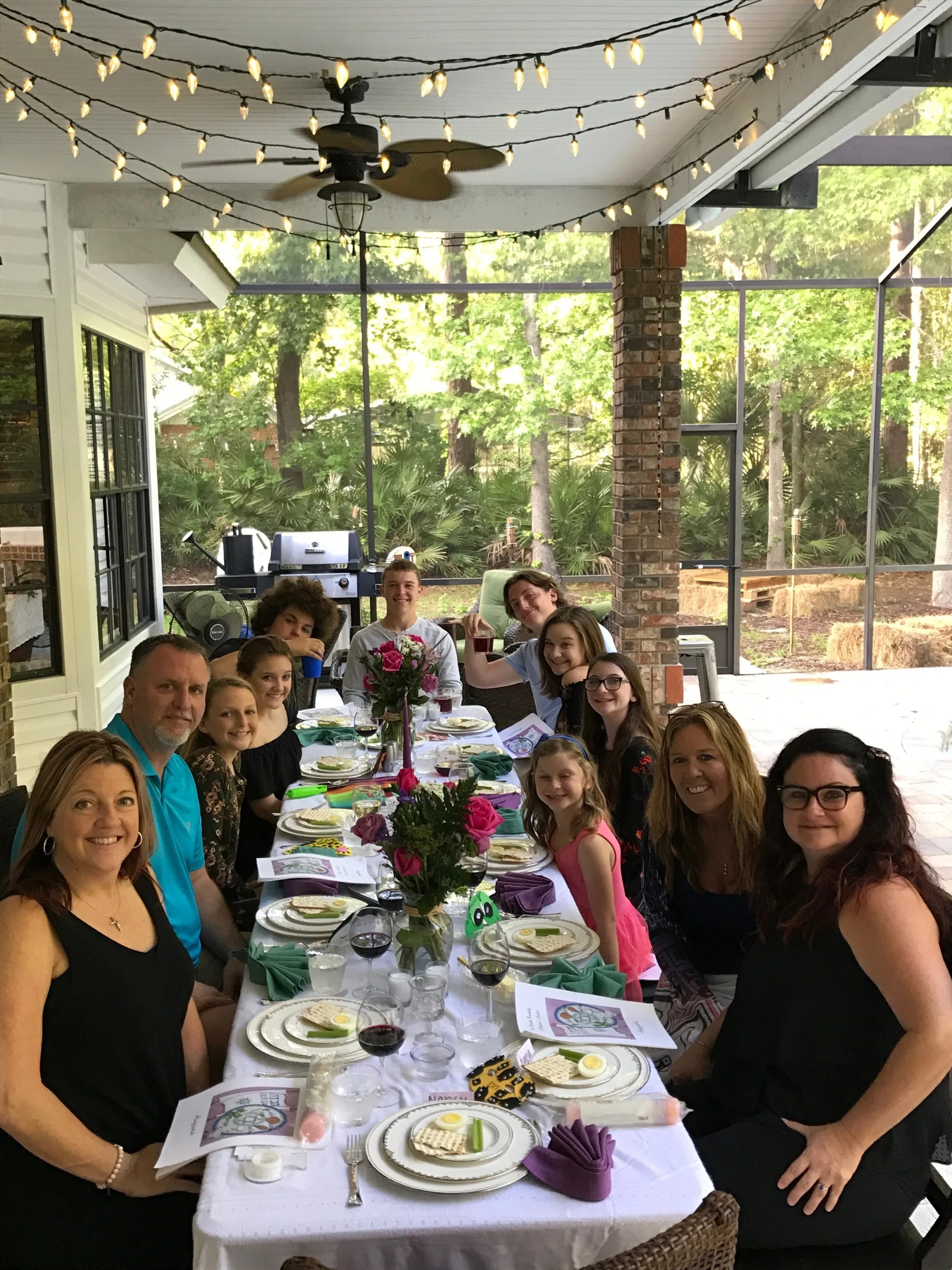 Family gathering with children and adults seated at a decorated outdoor dining table on a screened porch, with string lights overhead and a lush green background.