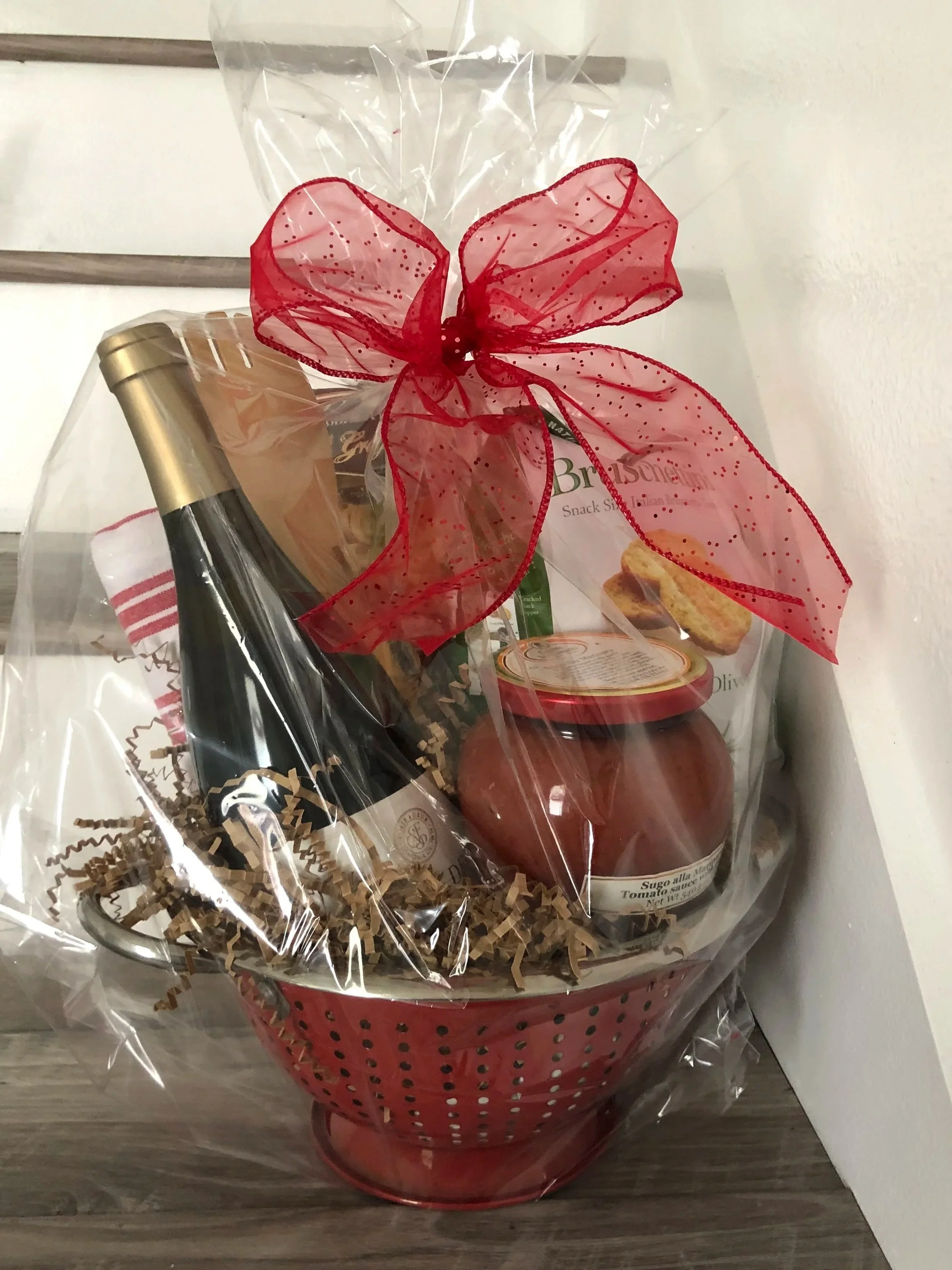 Gift basket wrapped in clear plastic with a large red bow, containing a bottle of wine, a jar of tomato sauce, and other food items, sitting on a wooden surface.