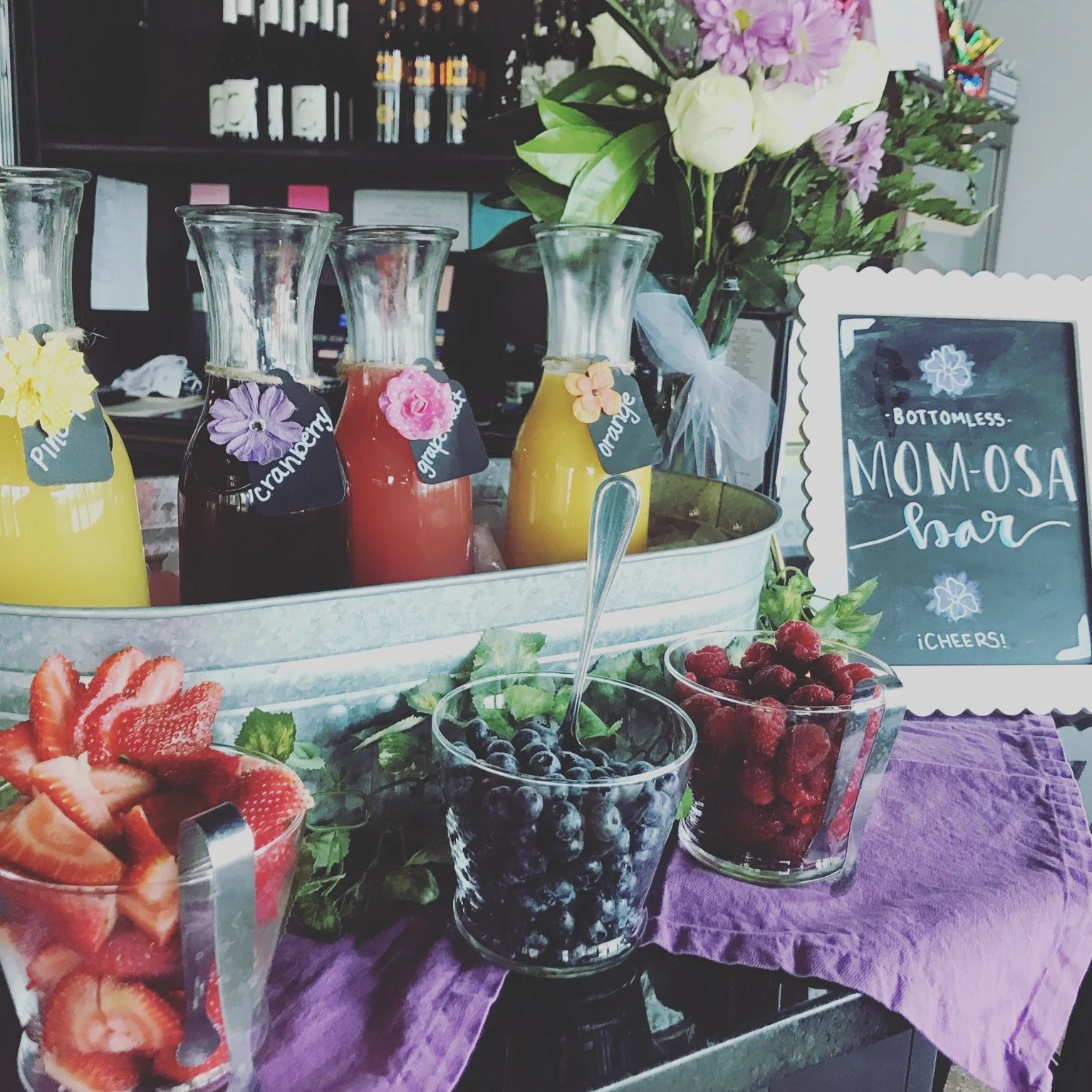 A display of colorful fruit smoothies in glass bottles, with side bowls of strawberries, blueberries, and raspberries, on a table with a purple cloth. There are fresh flowers and a chalkboard sign reading 'Bottomless MOM-OSA bar, Cheers!', at a brunc