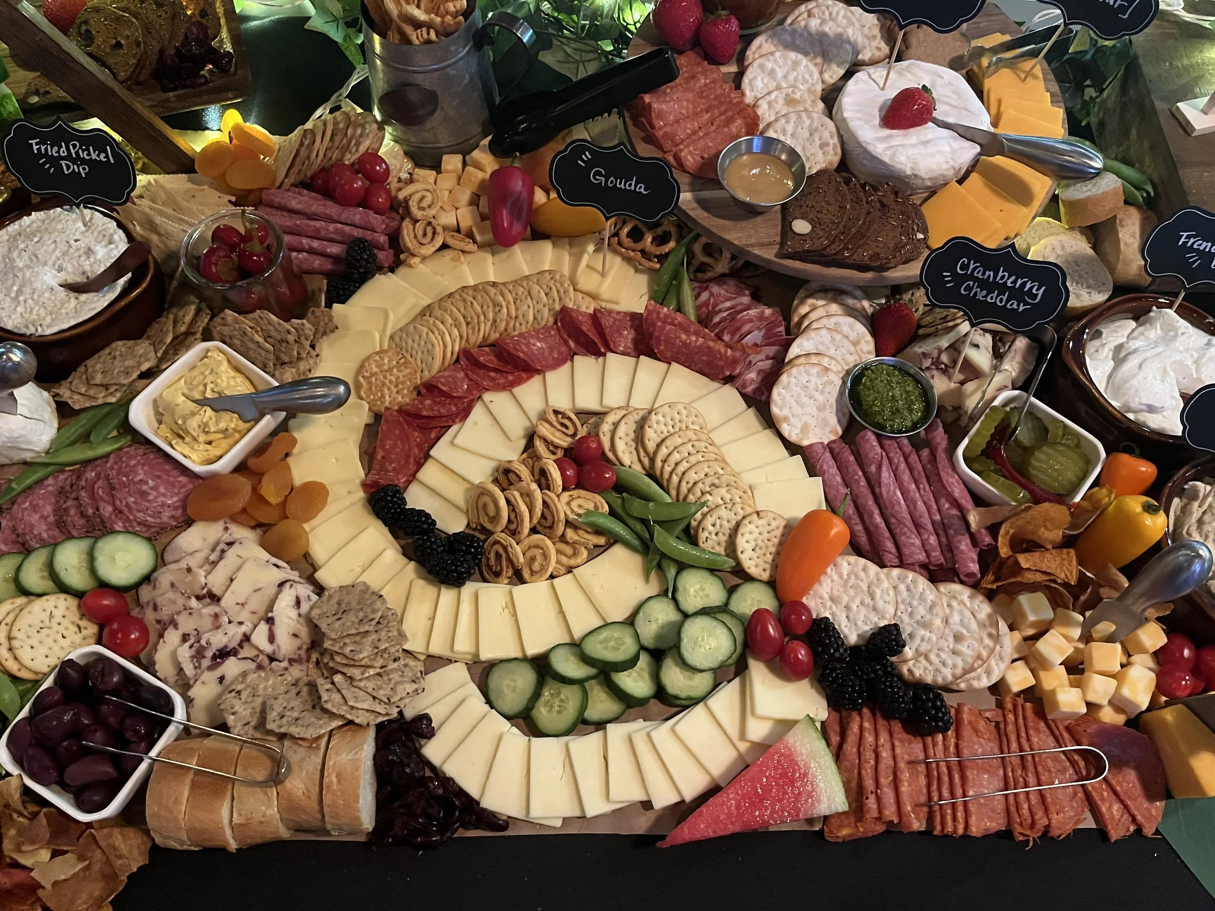 A large charcuterie board featuring various cheeses, cured meats, crackers, fruits, berries, pickles, and spreads, with small chalkboard labels identifying some of the items.