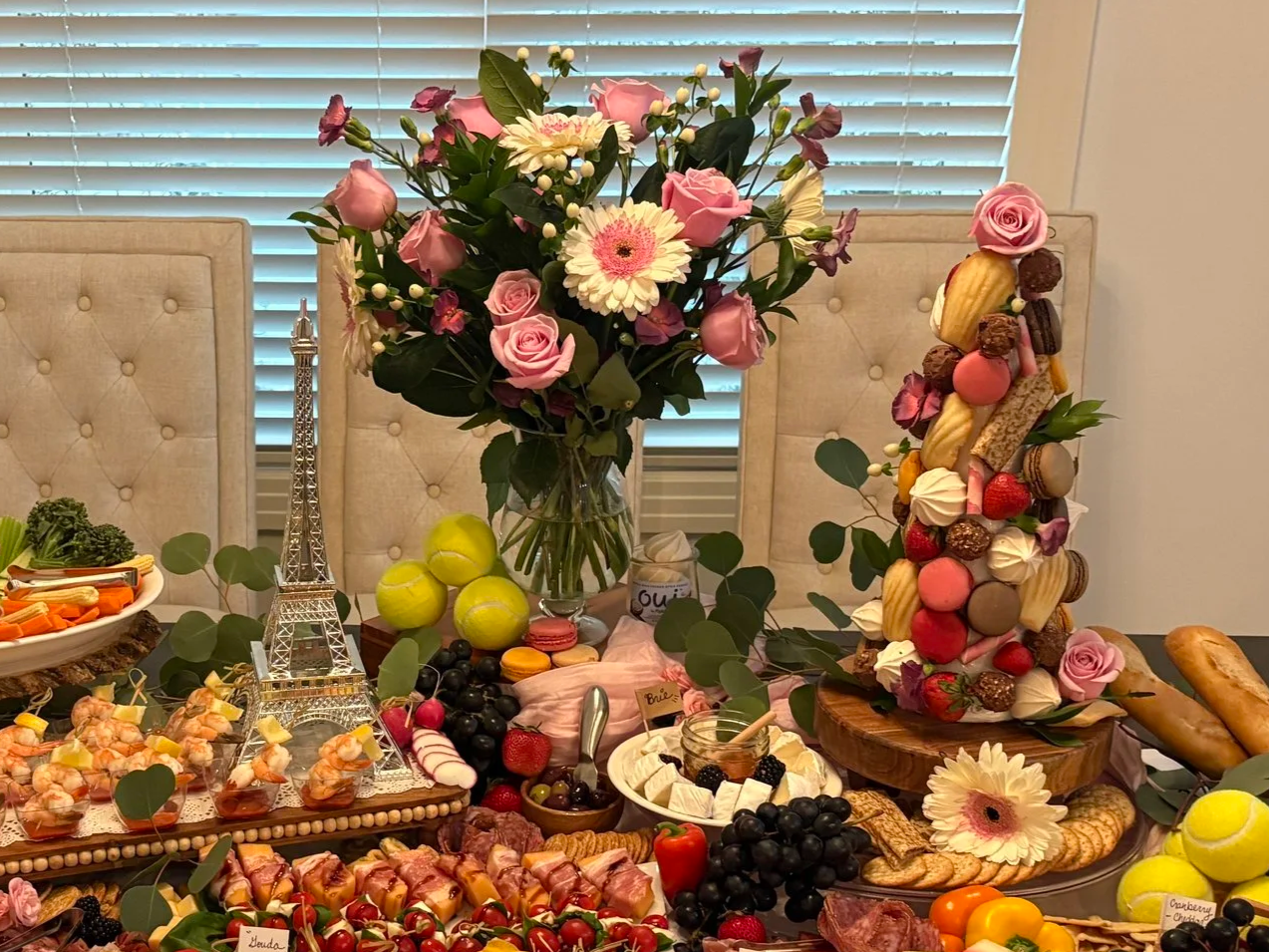 A table decorated with a large flower arrangement in a vase, an Eiffel Tower model, various desserts including macarons, strawberries, grapes, cheese, and baked goods, with a large rosemary and fruit centerpiece and a backdrop of window blinds.