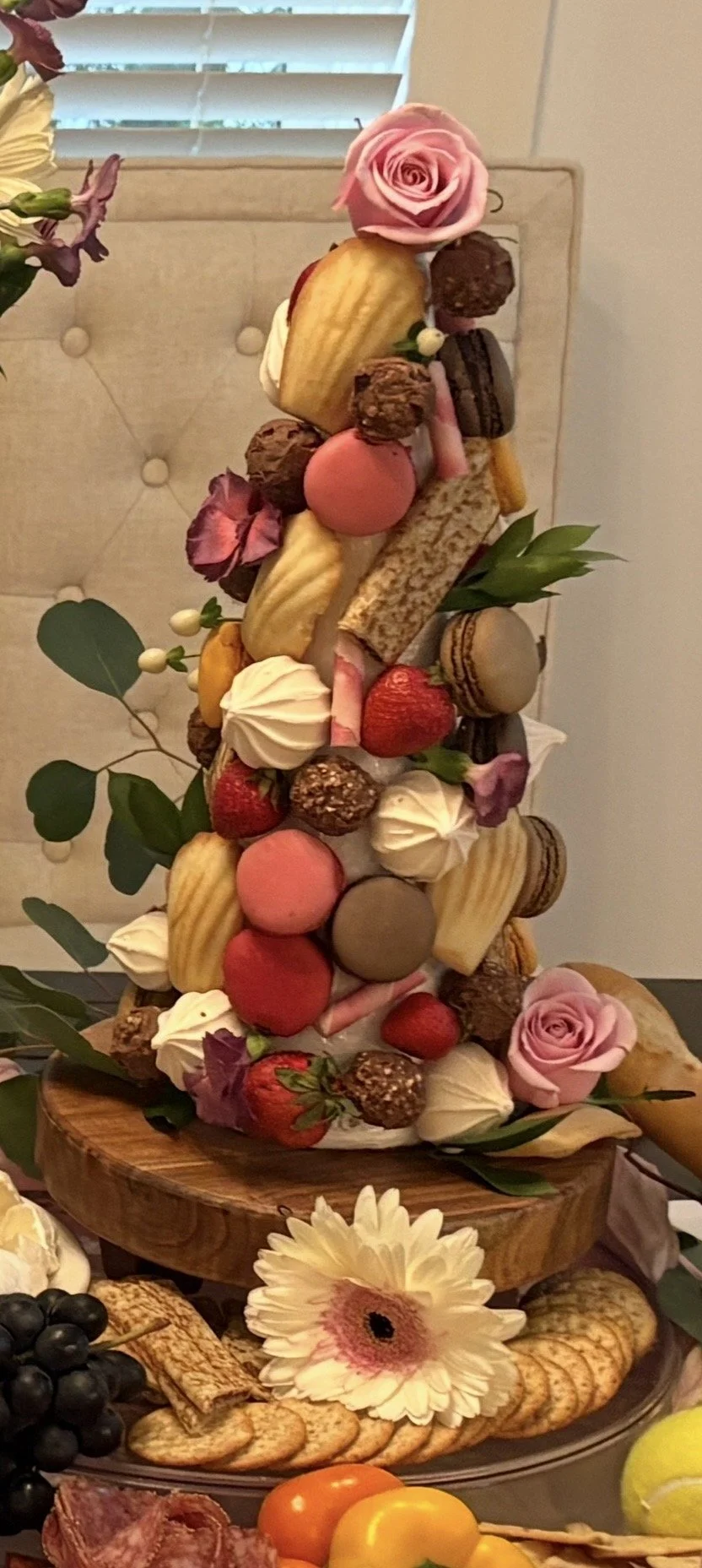 A tall dessert arrangement with macarons, strawberries, chocolates, flowers, and green leaves on a wooden base.