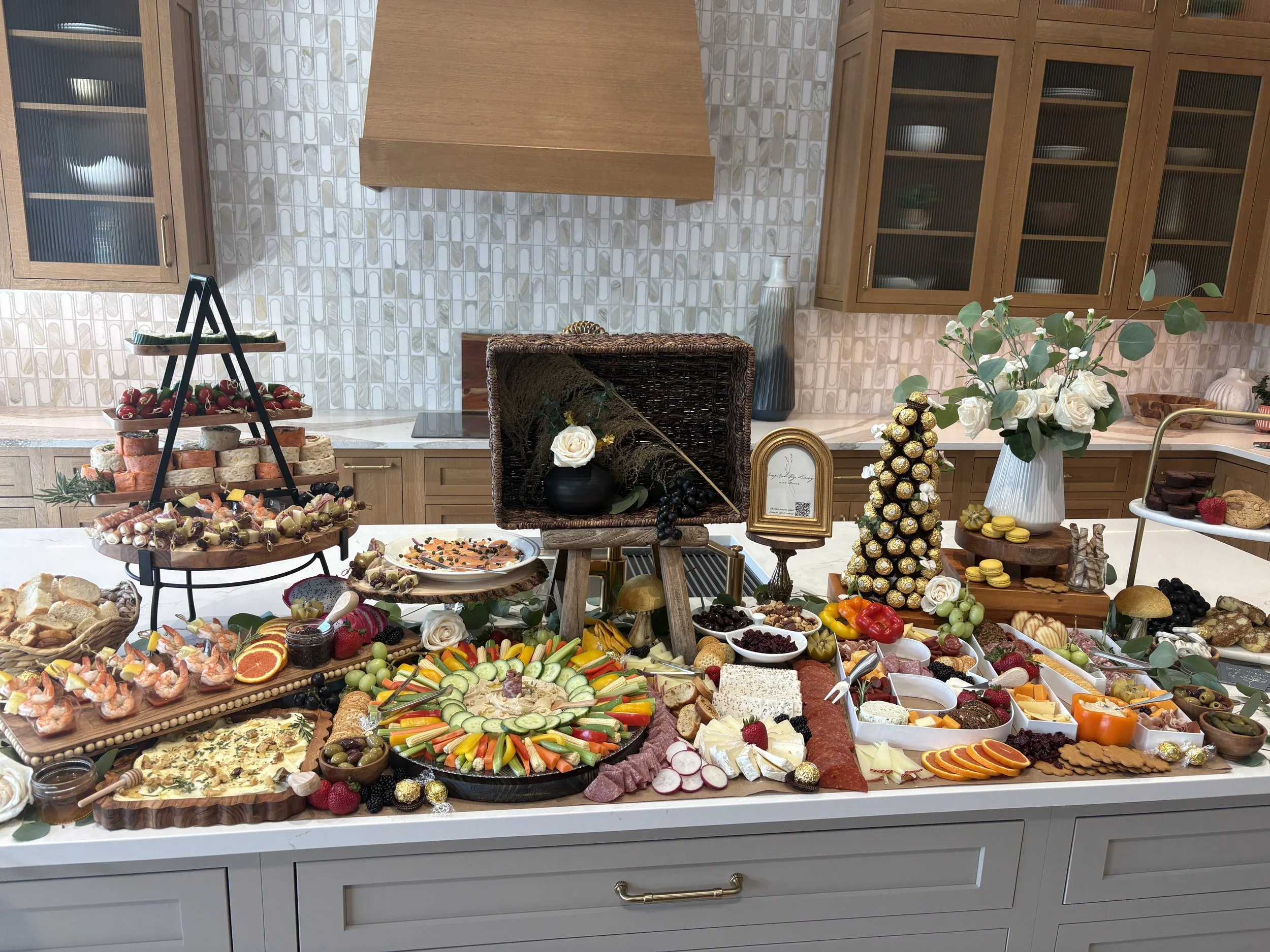 A kitchen buffet table filled with various finger foods, cheeses, fruits, vegetables, and desserts. There are also decorative flowers in a vase and small treats like chocolates and macarons. A large wooden cabinet is in the background.