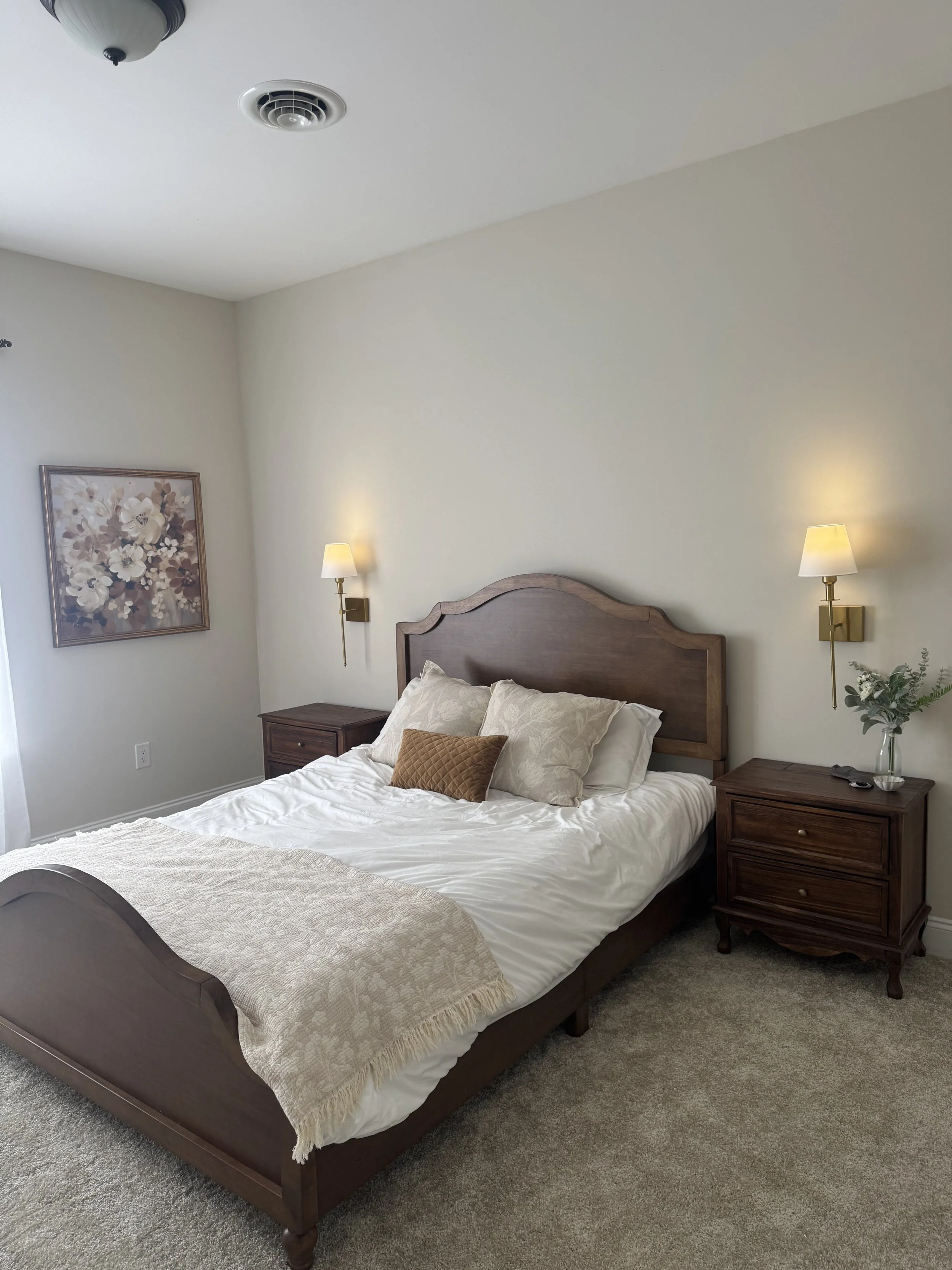 A bedroom with a wooden bed, beige pillows, a throw blanket, two matching nightstands with lamps, a vase with greenery, a floral painting, and a ceiling light fixture.