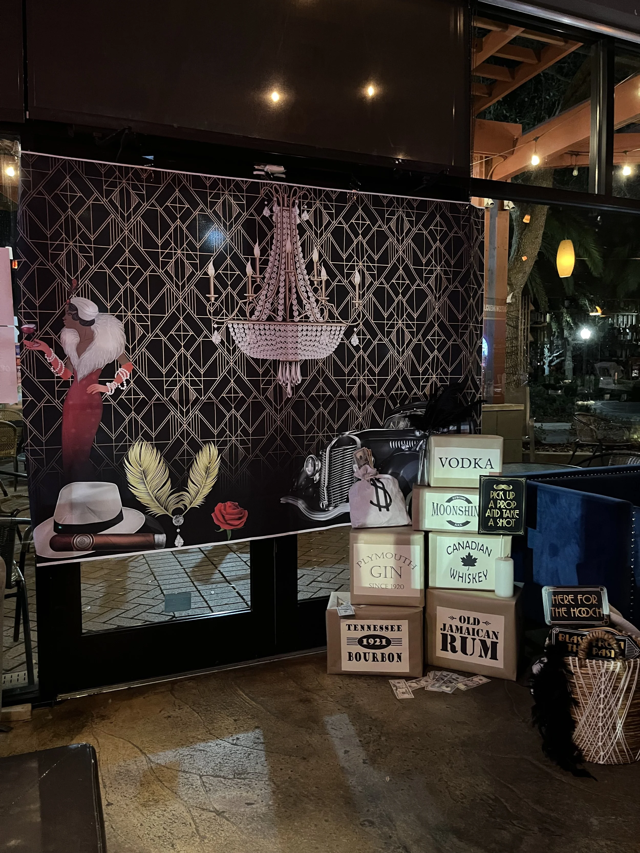 Decorative display featuring a black wall with a chandelier illustration, a vintage car illustration, a woman in a dress and fur stole, and a collection of illuminated boxes with labels for alcohol brands like vodka, moonshine, whiskey, gin, bourbon,