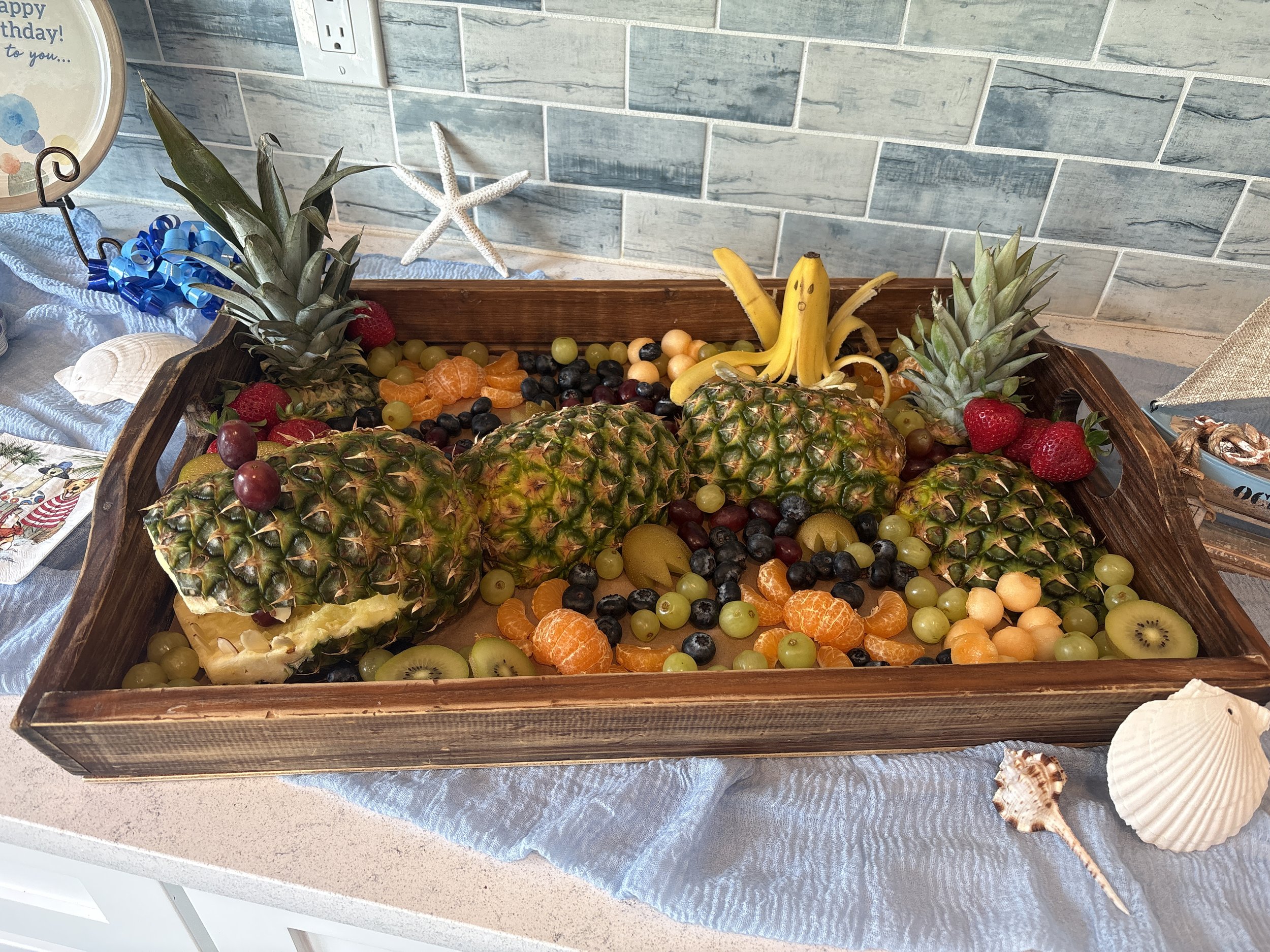 A wooden tray filled with a variety of fresh fruits including pineapples, strawberries, black grapes, green grapes, bananas, oranges, kiwis, and other mixed fruits, arranged on a countertop with a blue cloth underneath.
