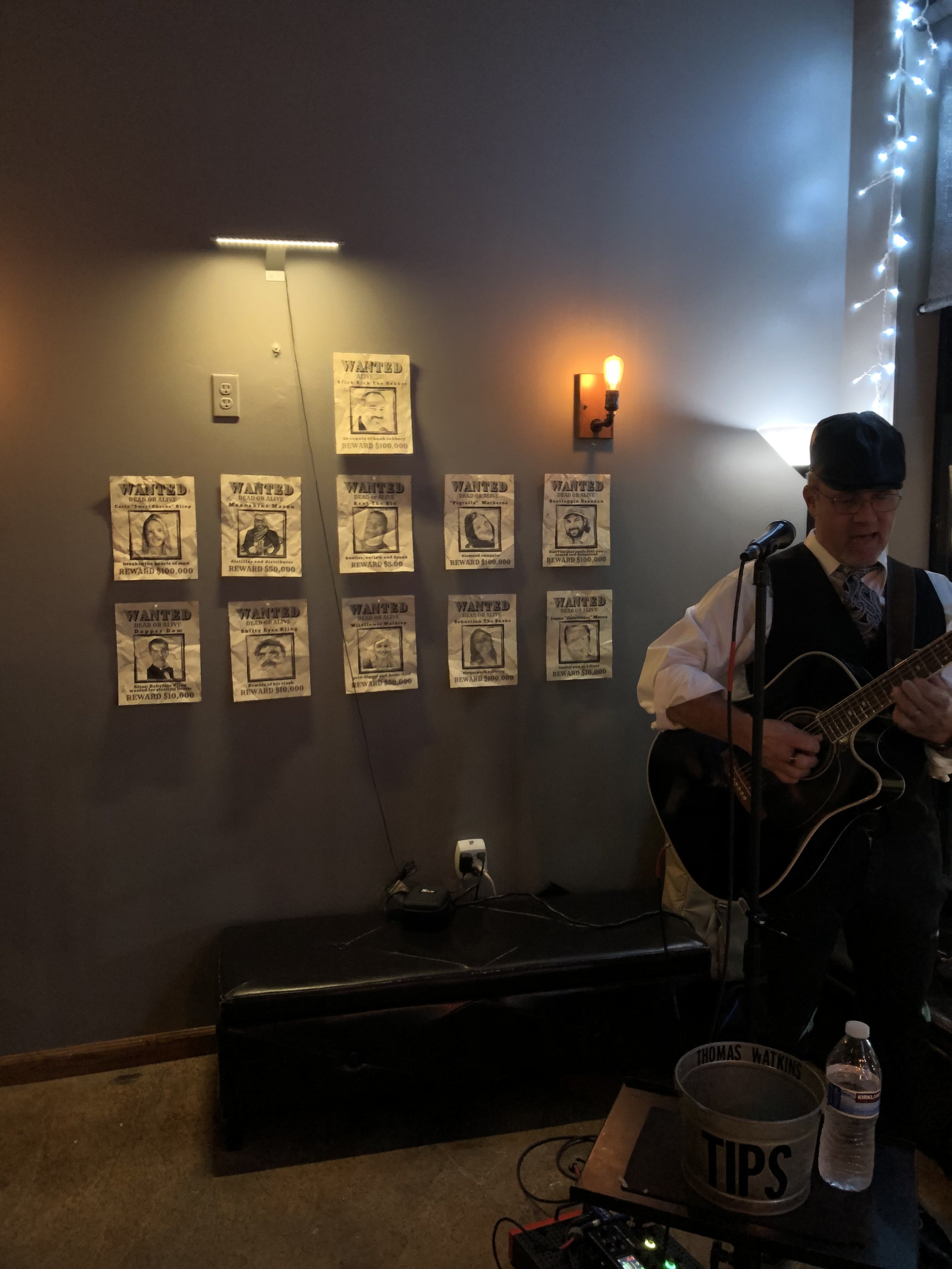 A man in vintage attire playing an acoustic guitar and singing in a dimly lit room. The wall behind him features a series of displayed wanted posters. There is a small lamp mounted on the wall and string lights in the corner.