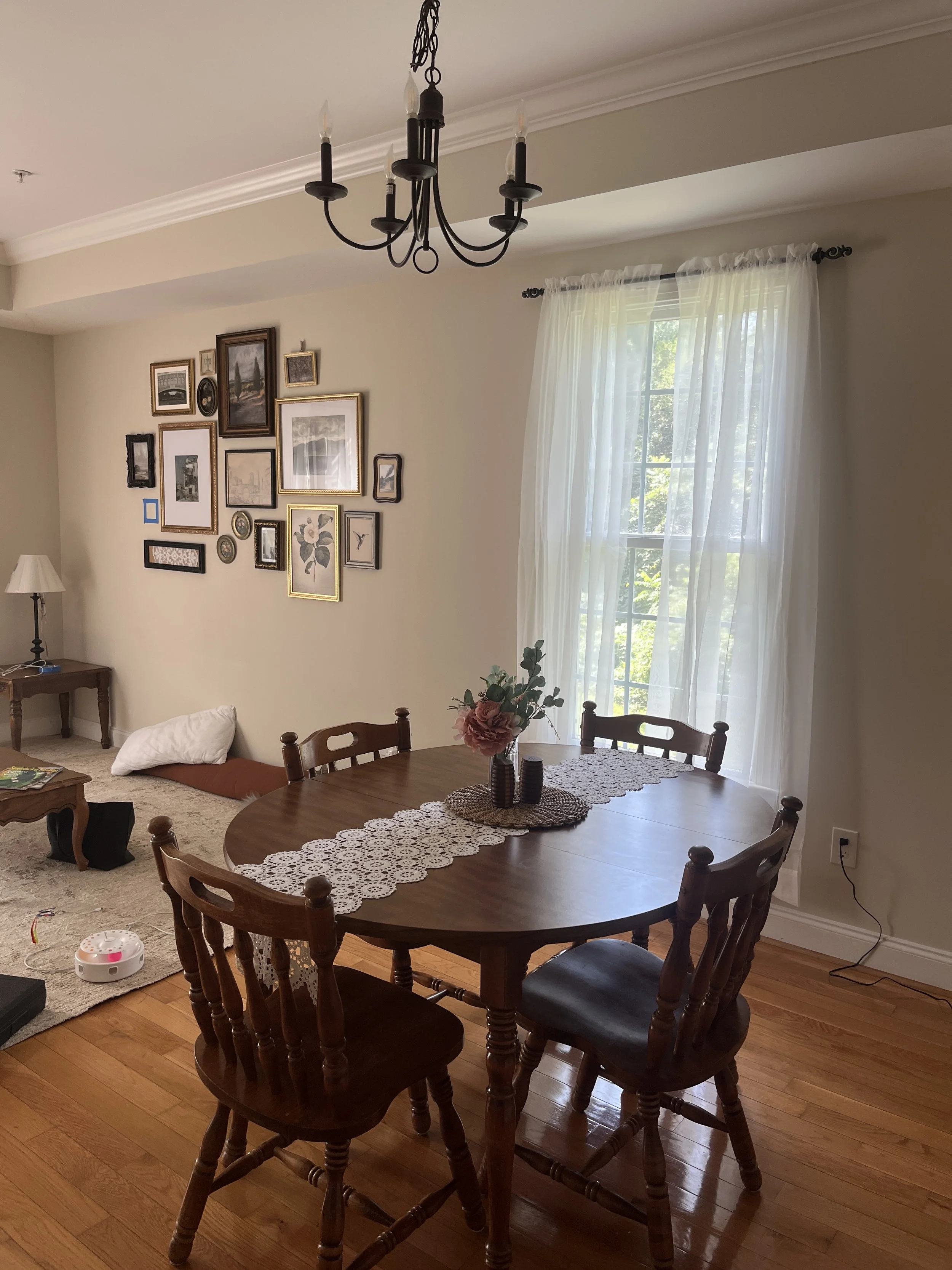 A dining room with a wooden table, four matching chairs, a window with sheer curtains, a collage of picture frames on the wall, a chandelier hanging from the ceiling, and a corner with a pillow and a small side table.
