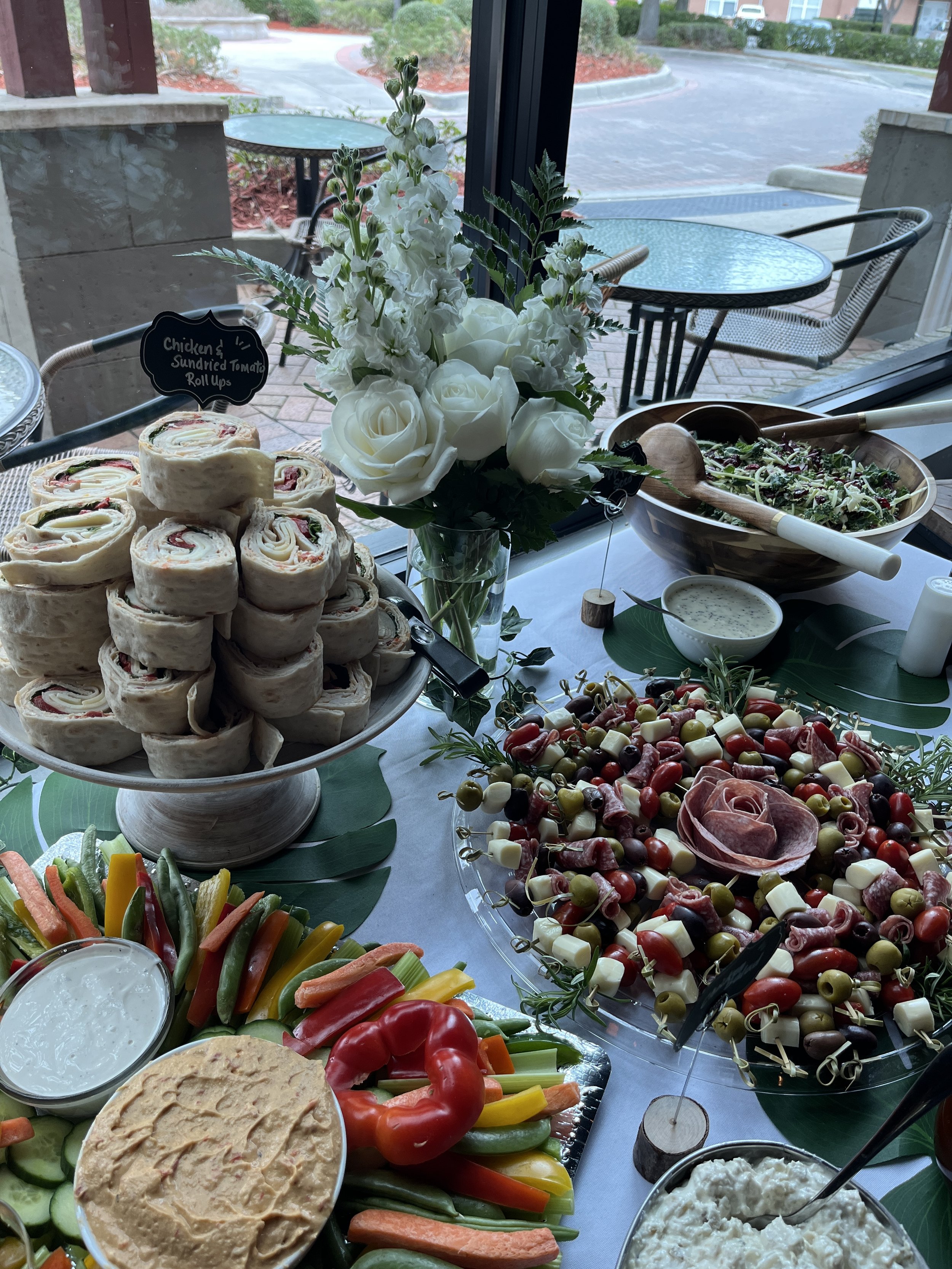 Vegetable platter with assorted sliced raw vegetables, cheese and meat skewers, a bowl of dip, a mixed salad, and a tray of rolled sandwiches on a table near a window with outdoor seating.