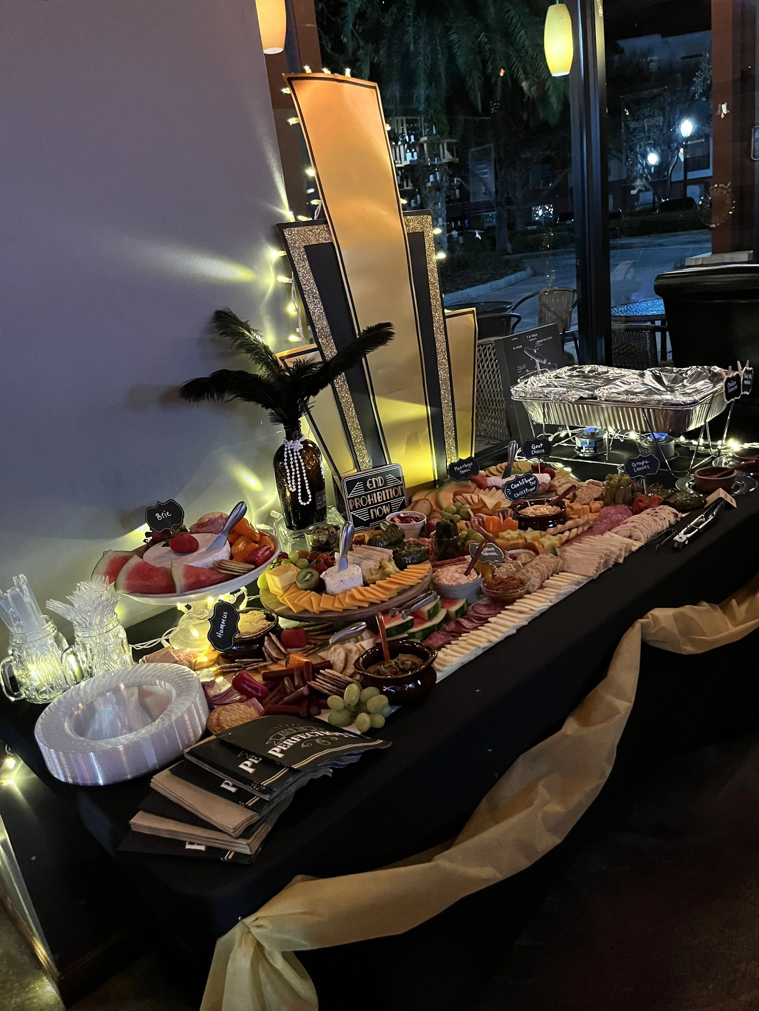 A decorated party buffet table with various fruits, meats, cheeses, crackers, and dips, illuminated by string lights and evening lighting, with a black tablecloth and elegant black, gold, and glittery decorations.