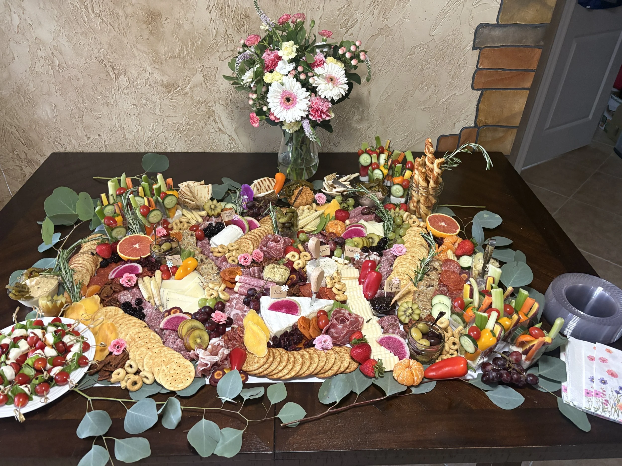 A table with a variety of food including colorful vegetables, cheeses, dips, crackers, and fruits, topped with a bouquet of pink and white flowers in a glass vase, and garnished with eucalyptus leaves.