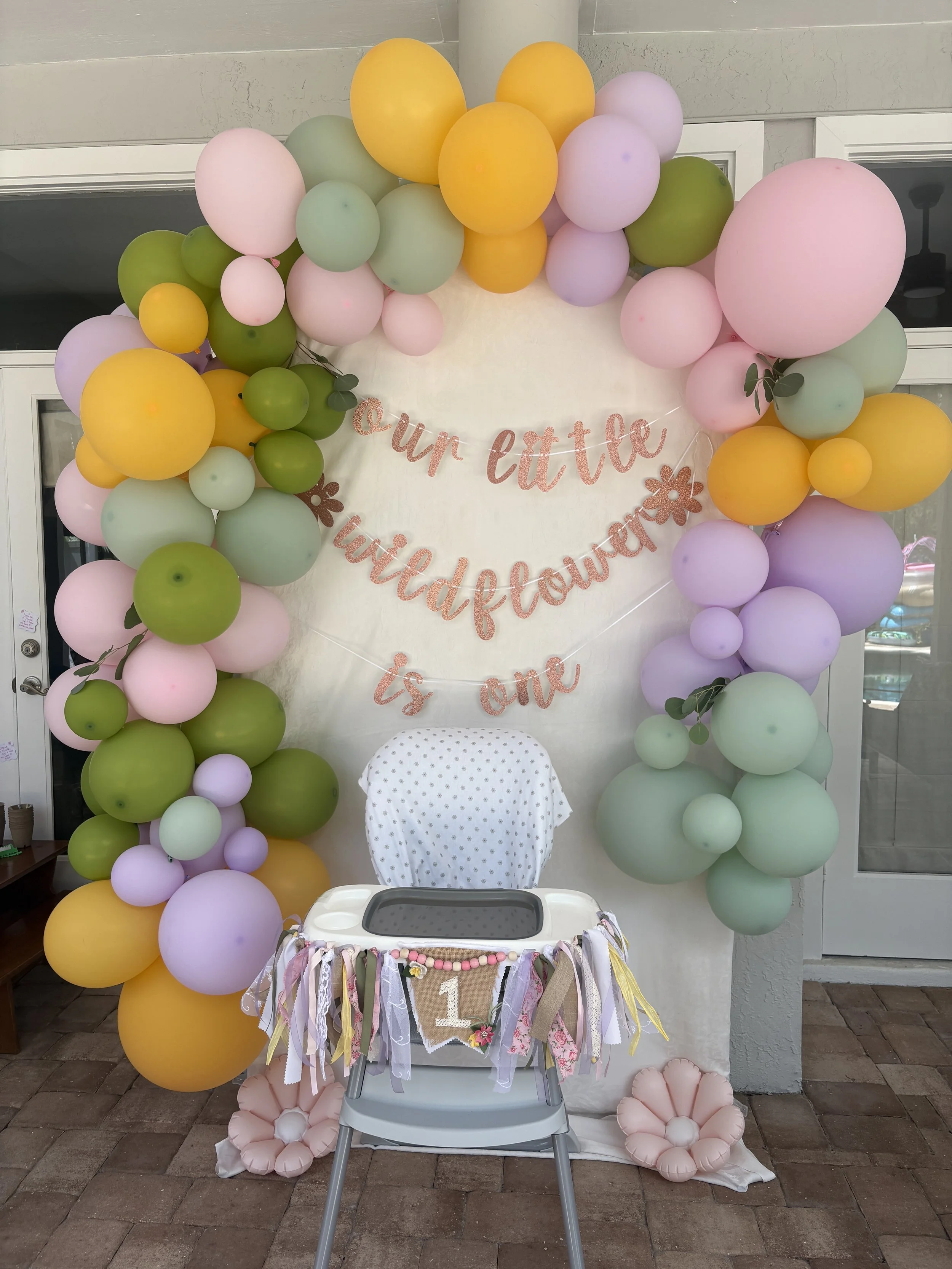 Decorative balloon arch with pink, green, yellow, and purple balloons surrounding a white backdrop that has a pink glittery banner reading '@pun ettk'. A small chair decorated with pastel ribbons and flowers is in front of the backdrop, and a beige a