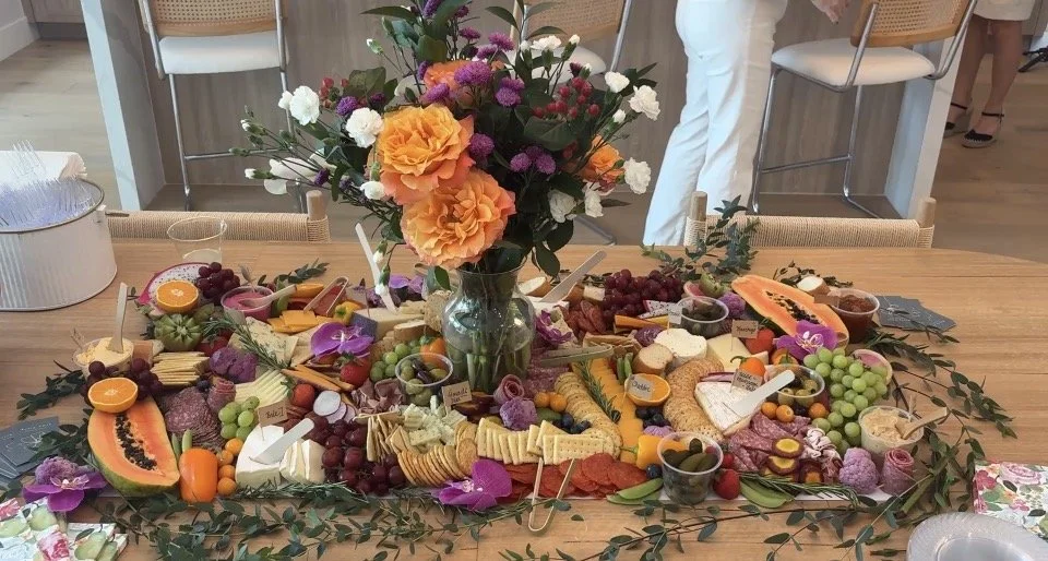 A table with a large floral centerpiece surrounded by an assortment of cheeses, fruits, nuts, and snacks, with some labels and small bowls on a party or gathering setting.