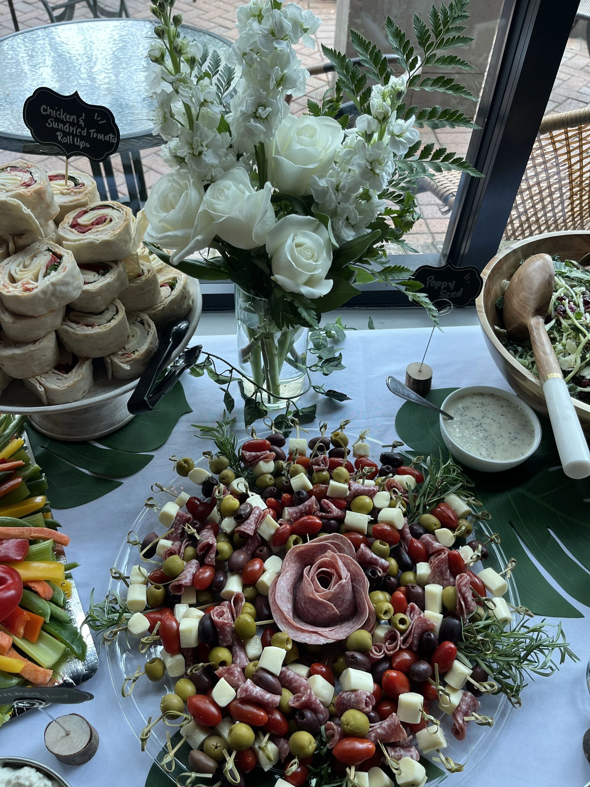 A table of appetizers featuring a charcuterie platter with cheese, cured meats, cherry tomatoes, and olives, a bowl of ranch dressing, and a veggie platter with sliced carrots, celery, and bell peppers. There is a large floral arrangement with white 