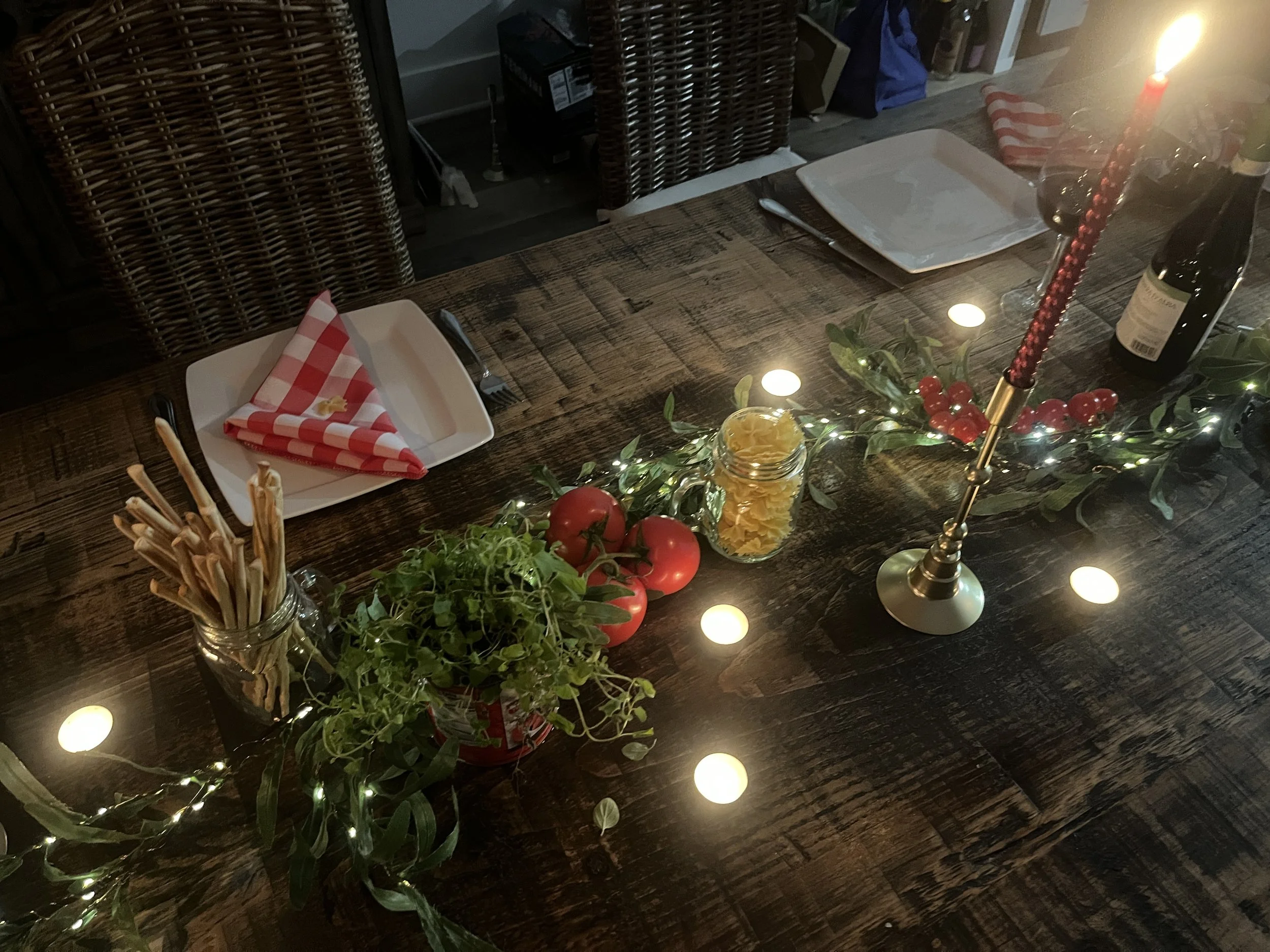 A cozy dining table decorated for Christmas with a candle centerpiece, fairy lights, tomatoes, a jar of pasta, and place settings with red and white napkins.