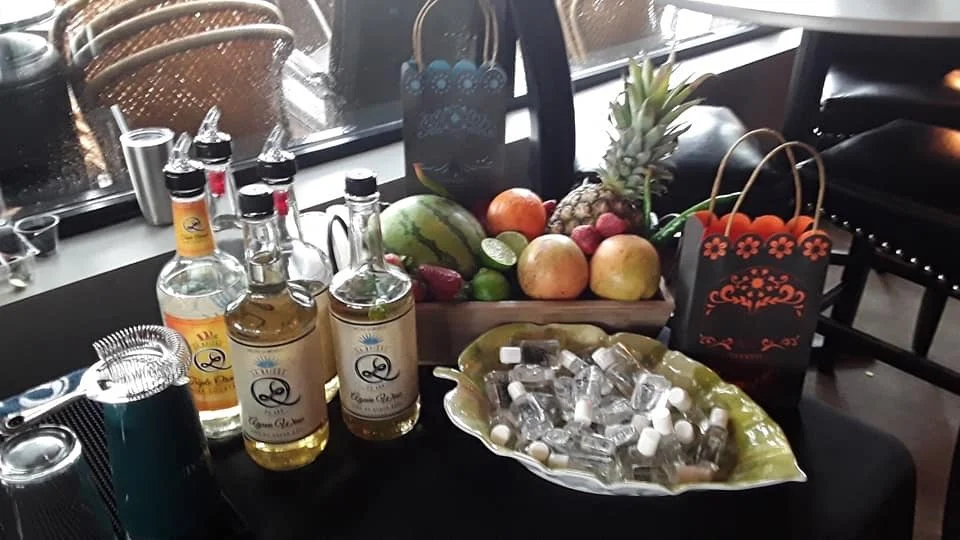 Table with bottles of liquor, a basket of assorted fruits, a bowl of small bottles, and gift bags, set near a window.
