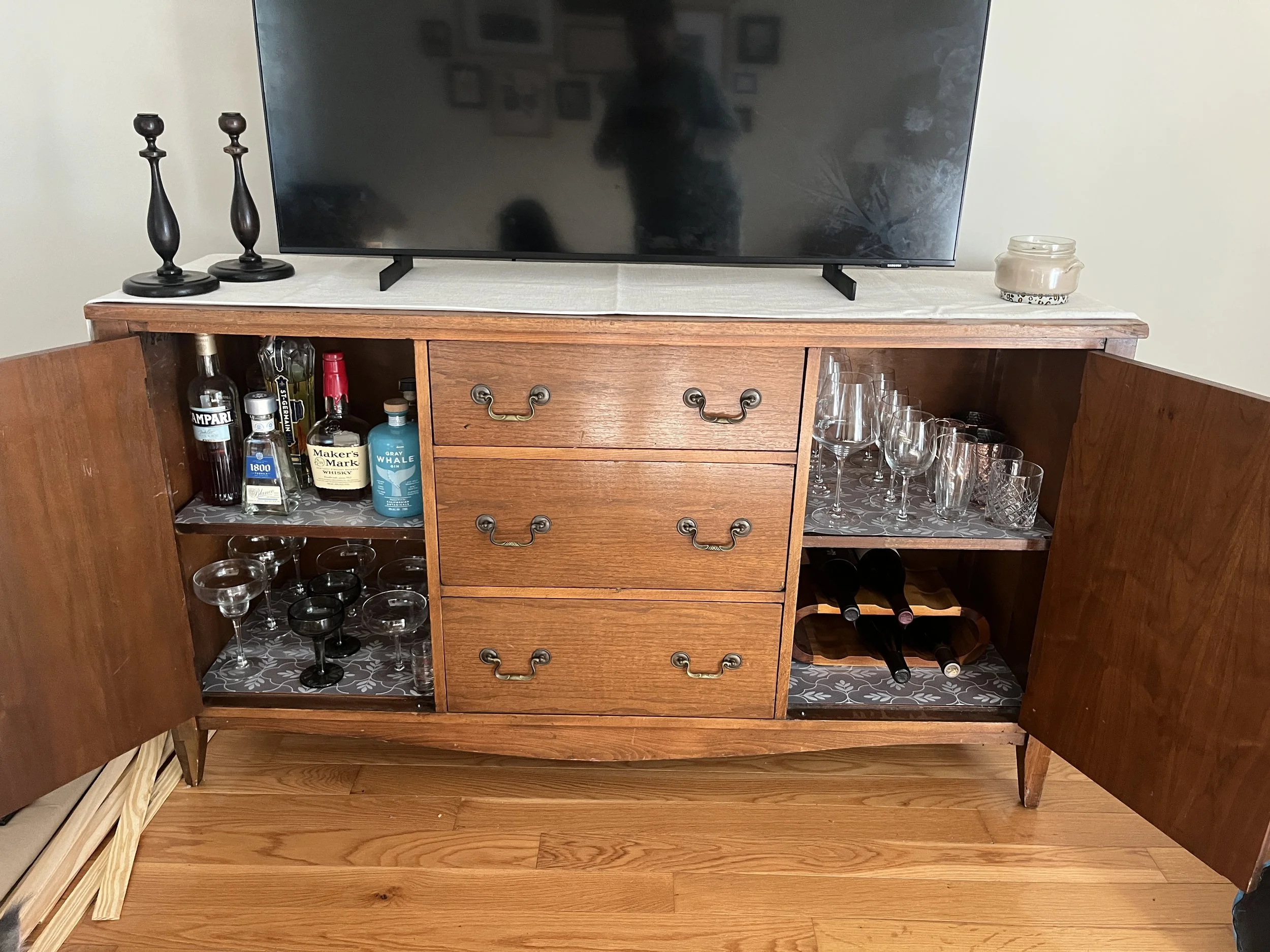 A vintage wooden bar cabinet with its doors open, displaying liquor bottles, glassware, and wine bottles inside. A television is on top of the cabinet, with candles and a decorative jar on either side.