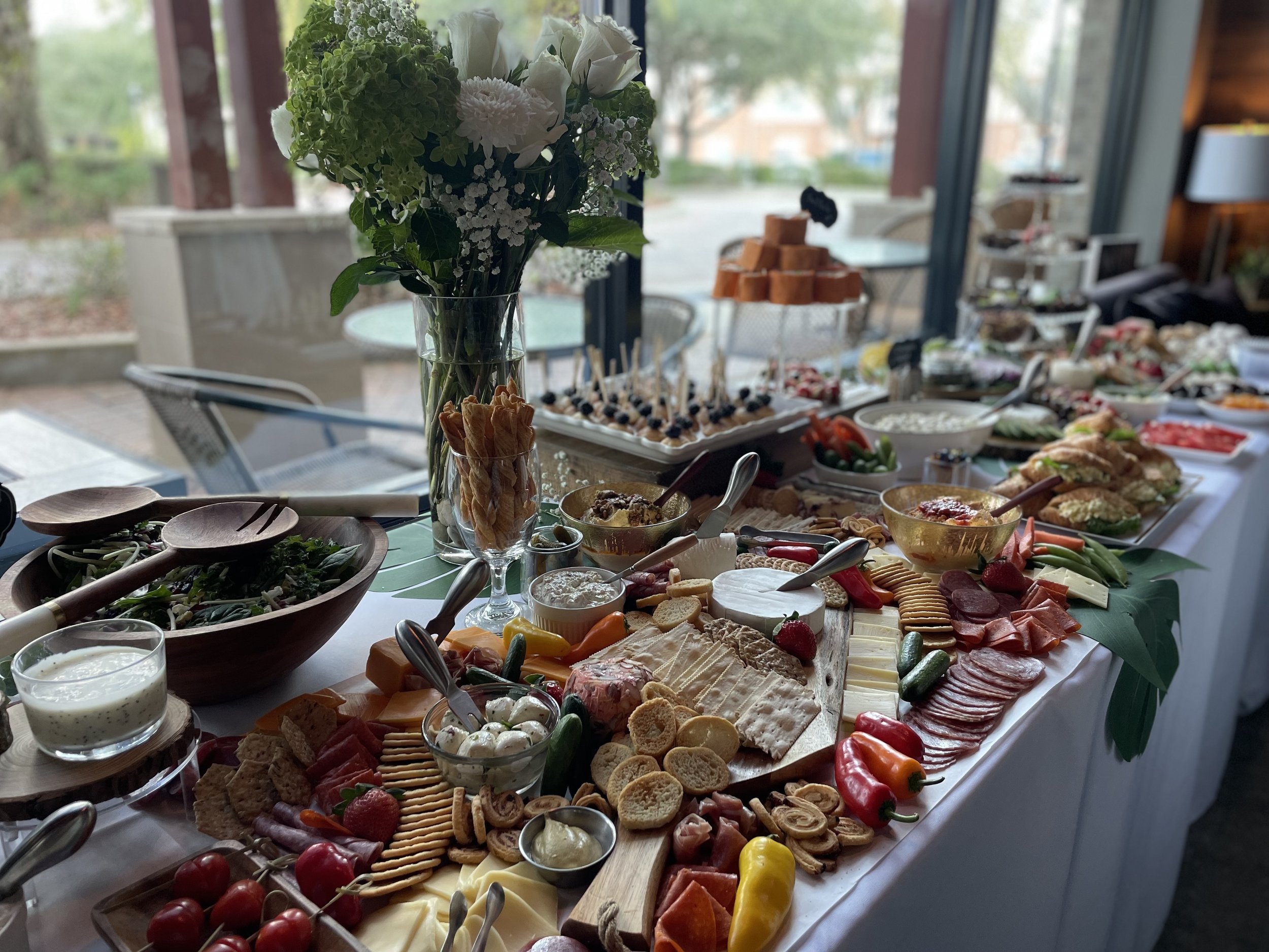 Buffet table with a variety of foods, including cheeses, meats, vegetables, fruit, crackers, dips, and desserts, with a vase of white flowers in the center.