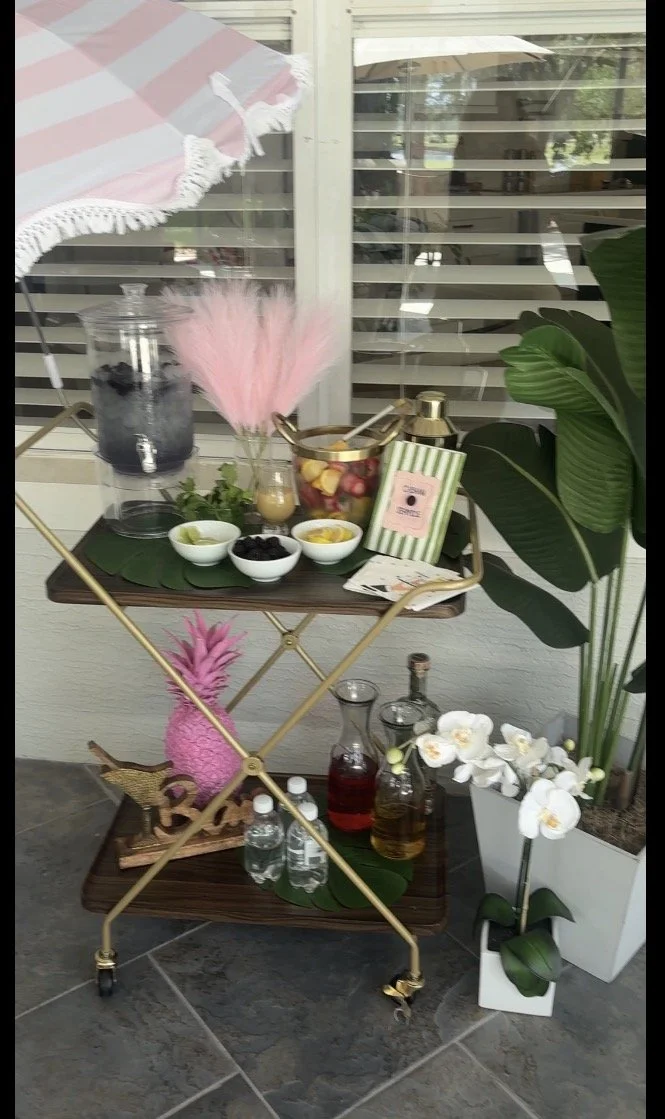 A gold and wood rolling cart with two shelves holding decorative items and beverages. The top shelf has small bowls of snacks, a pink feathered decoration, a container of apples, and a green striped package. The bottom shelf has three bottles of drin