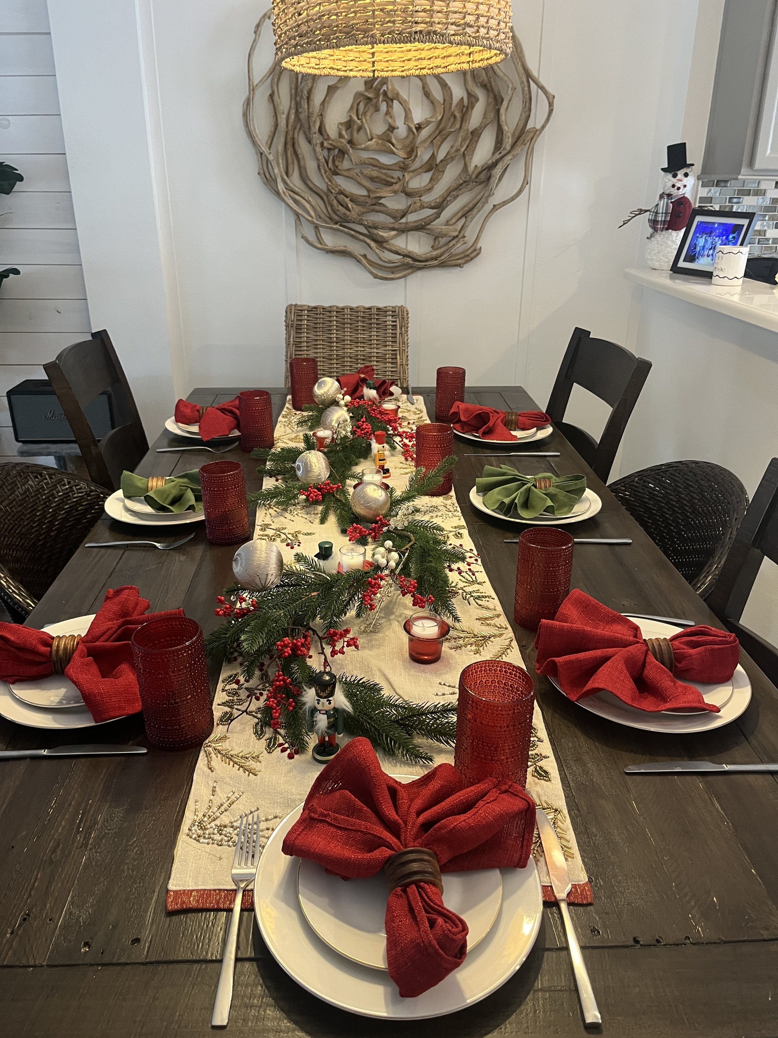 A festive Christmas dining table with red and green napkins, silver and red candles, and holiday decorations including pine branches, red berries, Christmas ornaments, and a nutcracker, set in a cozy room.
