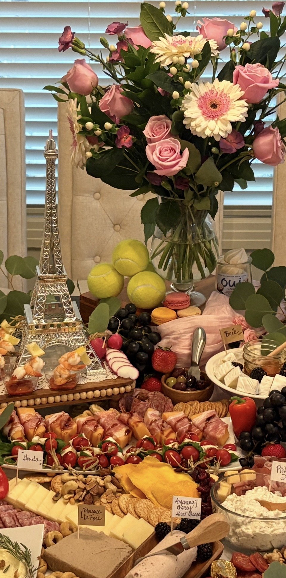 A table set with a large bouquet of pink roses and white gerbera daisies in a glass vase, surrounded by green leaves. The table also features tennis balls, a miniature Eiffel Tower, various fruits, cheeses, crackers, cured meats, macarons, and small 